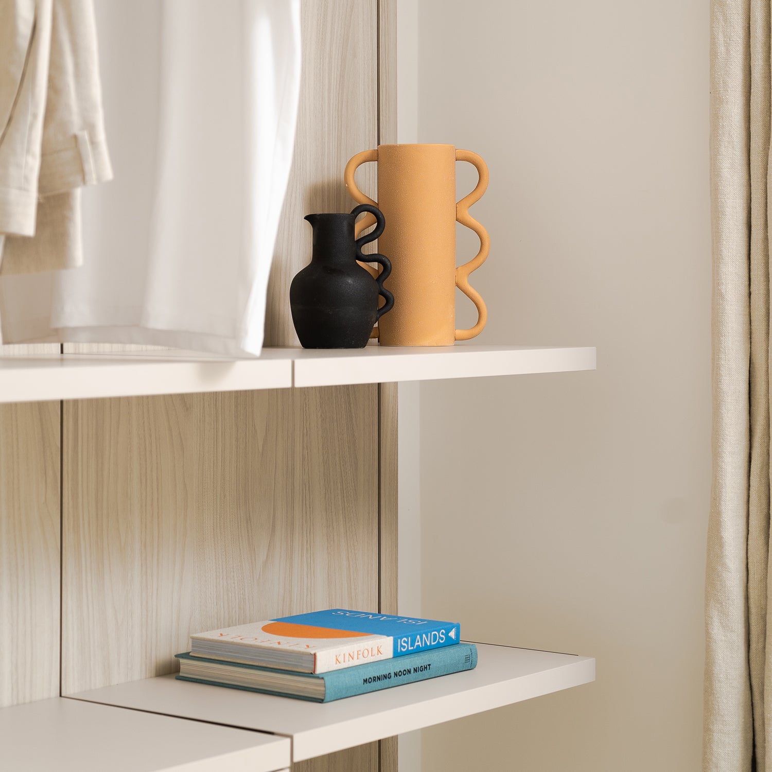 Contemporary wardrobe shelving with sculptural ceramic vases in black and terracotta, neatly folded clothing, and stacked lifestyle books including Kinfolk and Islands.