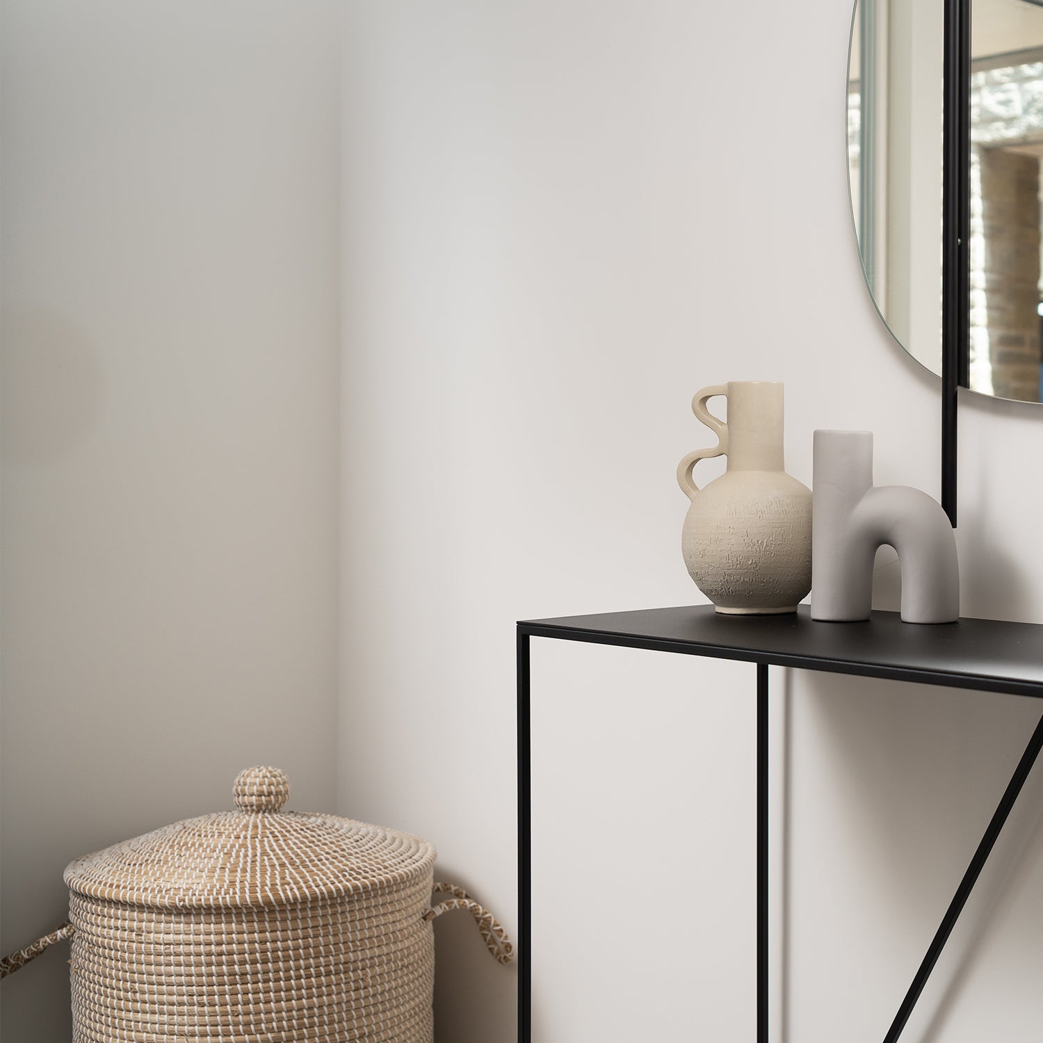 Minimalist hallway corner with black metal console table, sculptural ceramic vases, round woven basket with lid, and soft neutral walls reflecting Scandinavian-inspired interior styling.