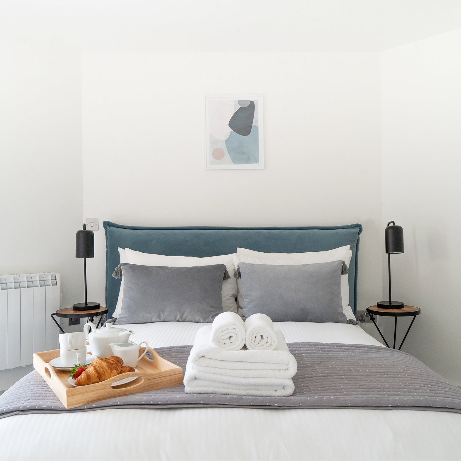 Stylish hotel bedroom interior by Iroka Interiors in Cornwall, showcasing a modern upholstered bed with plush grey cushions, bedside tables with black lamps, and a welcoming breakfast tray – ideal for boutique hospitality design inspiration.
