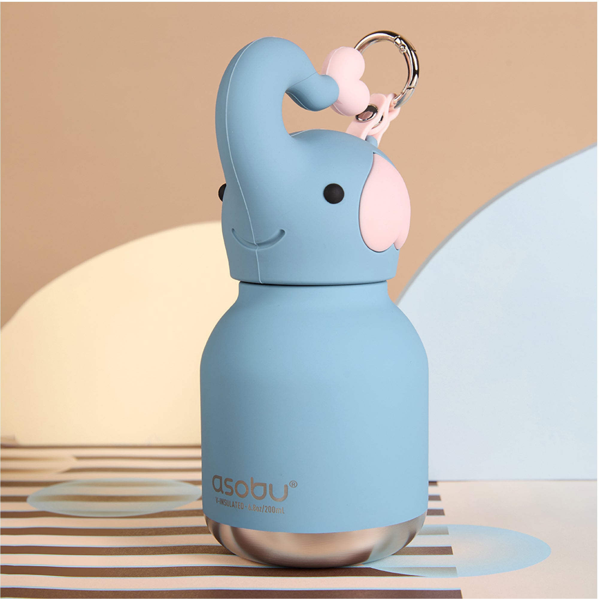 A light blue insulated water bottle shaped like an elephant, with a pink ear, black eyes, a smiling mouth, and a metal key ring attached. The bottle stands on a striped surface against a soft beige and blue studio background.