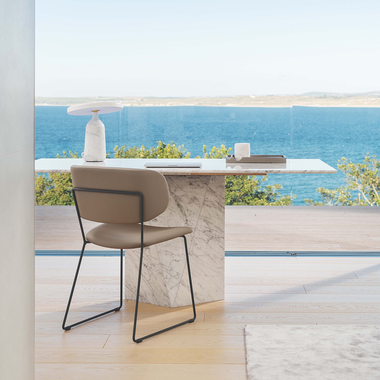 Minimalist home office with marble desk, taupe modern chair, and panoramic ocean view through floor-to-ceiling glass – cornish coastal workspace design with natural light and serene styling.