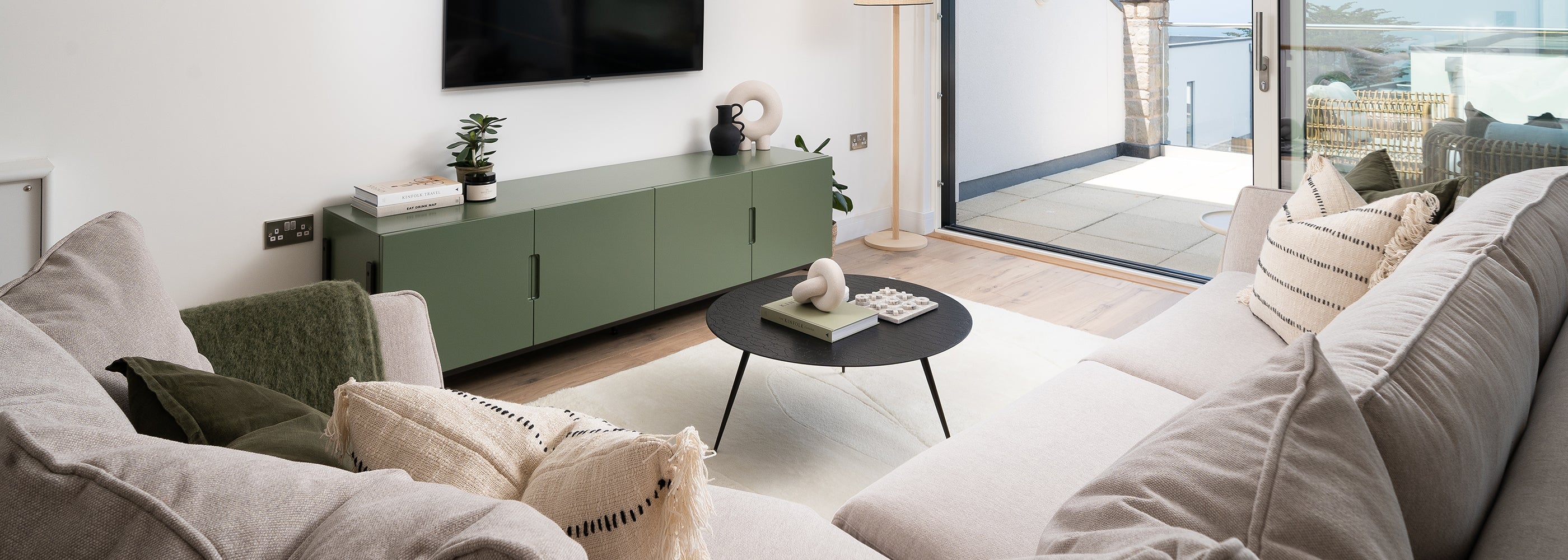 Modern coastal living room interior by Iroka Interiors in Hayle, Cornwall, featuring neutral sofas, olive green sideboard, black round coffee table, and sliding doors opening to a sea-view terrace – bespoke furniture and interior styling for contemporary Cornish homes.