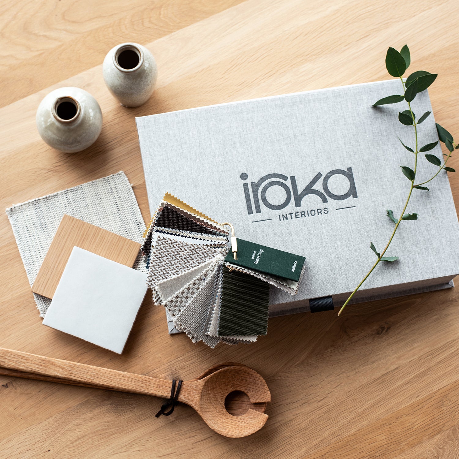 Iroka Interiors design kit with neutral fabric swatches, wood and tile samples, ceramic vases, and mood board elements on oak surface – curated interior design palette for modern home styling.