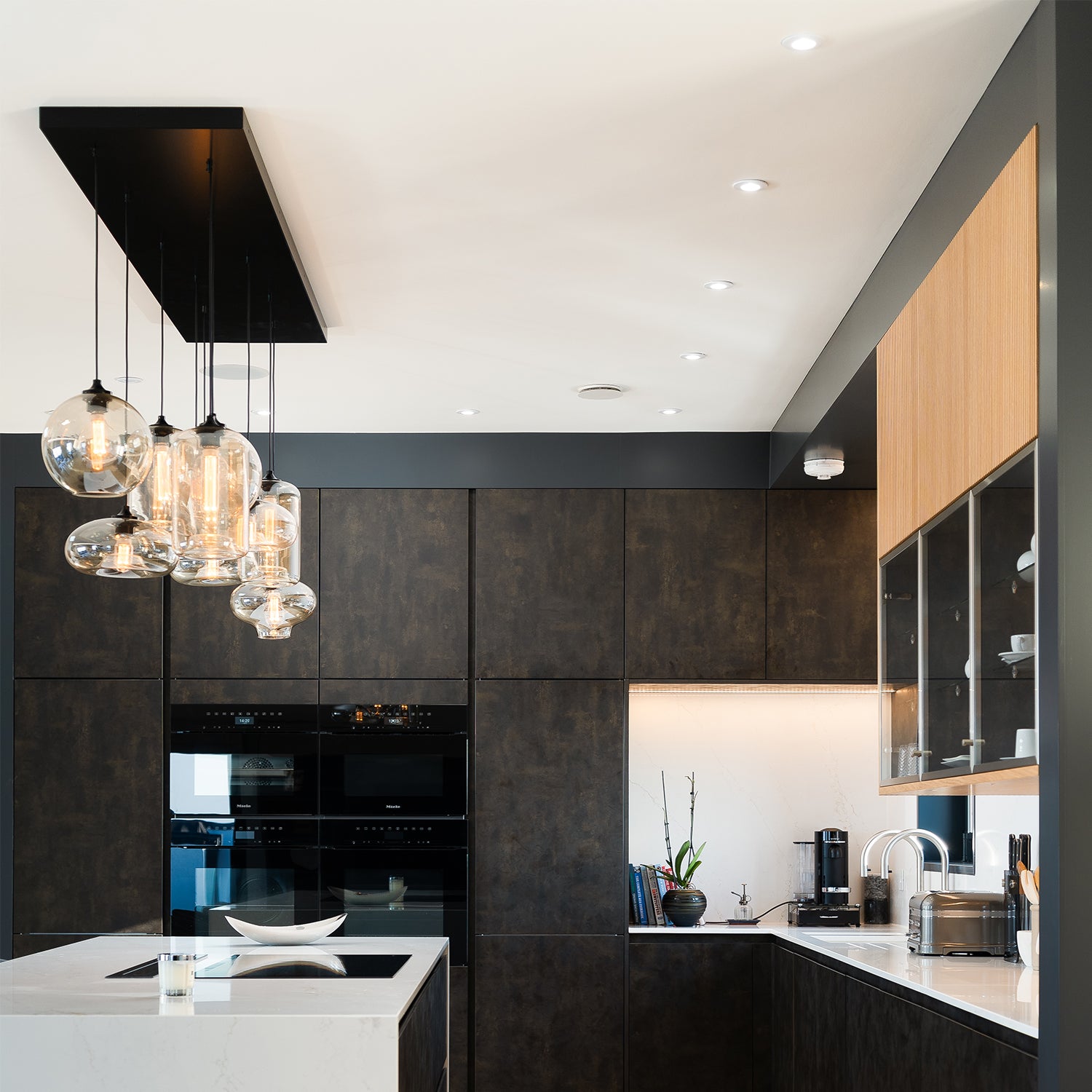"Luxury modern kitchen with dark textured cabinetry, warm wood accents, cascading glass pendant lighting, and a sleek white quartz island – custom kitchen design and installation by Iroka Interiors in Cornwall.