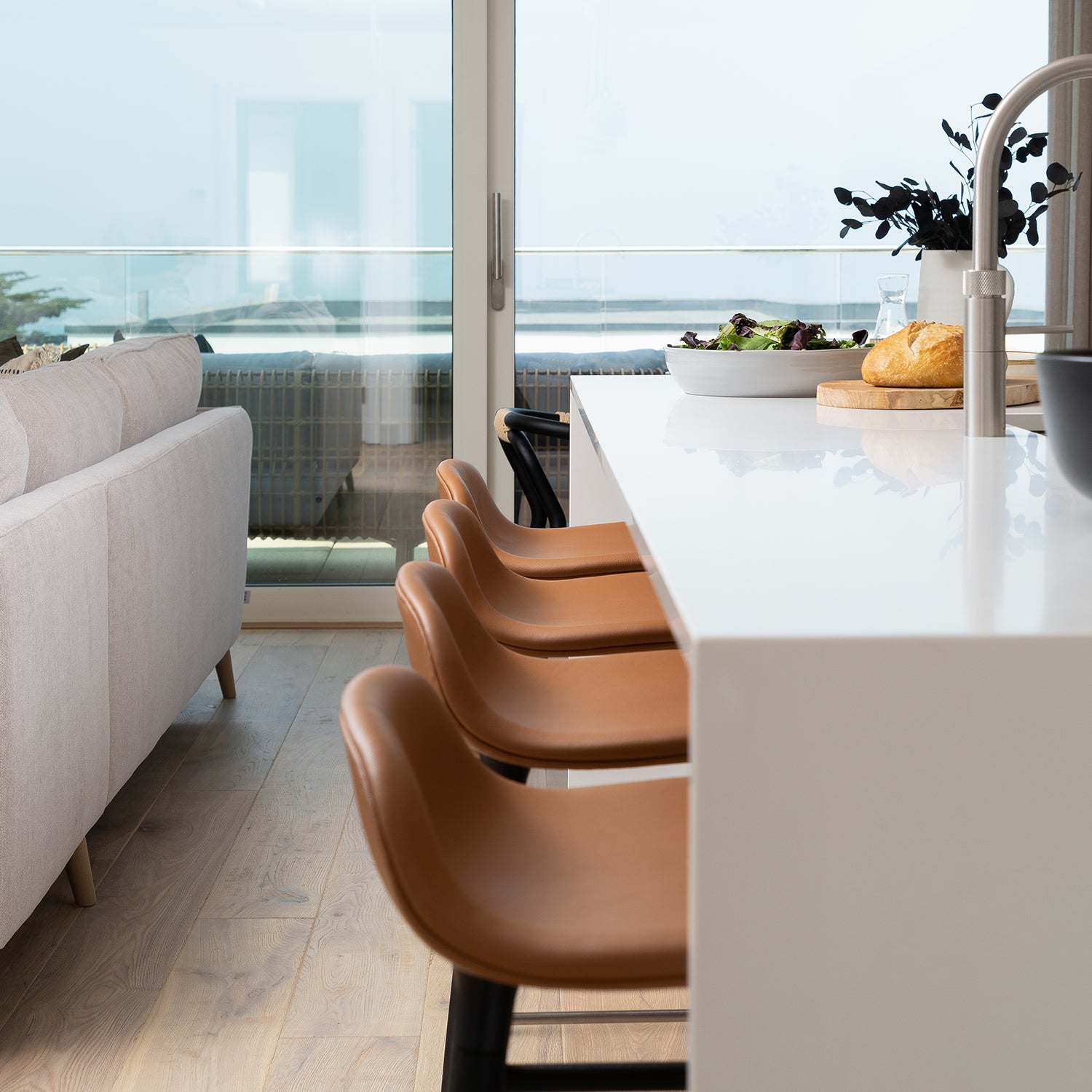 Contemporary coastal kitchen design by Iroka Interiors showcasing a sleek white waterfall island, tan leather bar stools, brushed steel tap, and panoramic sea views – bespoke kitchen installation in Cornwall.