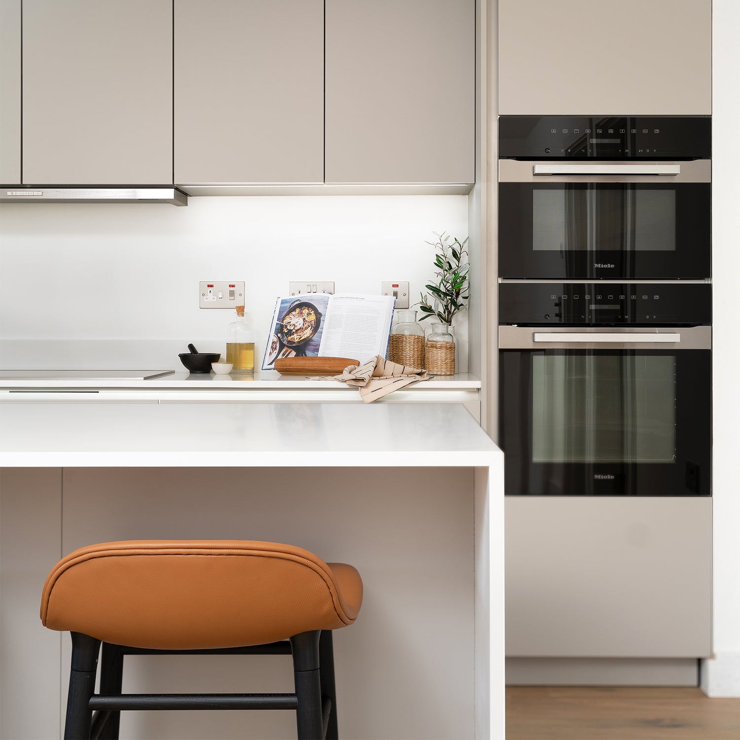 Modern kitchen design detail by Iroka Interiors featuring built-in Miele appliances, handleless taupe cabinetry, white quartz worktop with integrated hob, and warm tan leather barstool – bespoke kitchen installation in Cornwall.