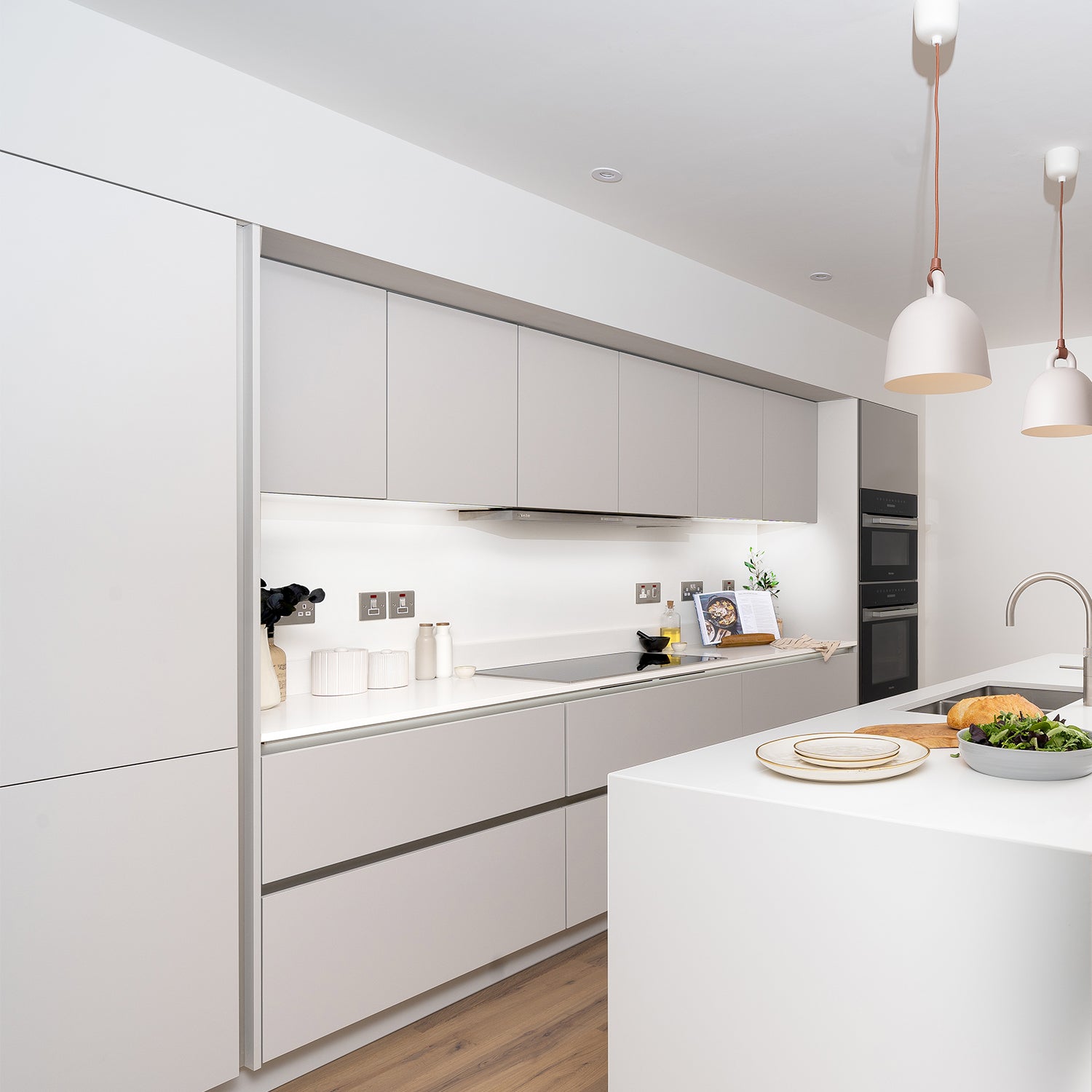 Contemporary white kitchen design by Iroka Interiors showcasing sleek handleless cabinets, curved island with integrated hob, minimalist lighting, and seamless built-in appliances – bespoke kitchen installation in Cornwall.