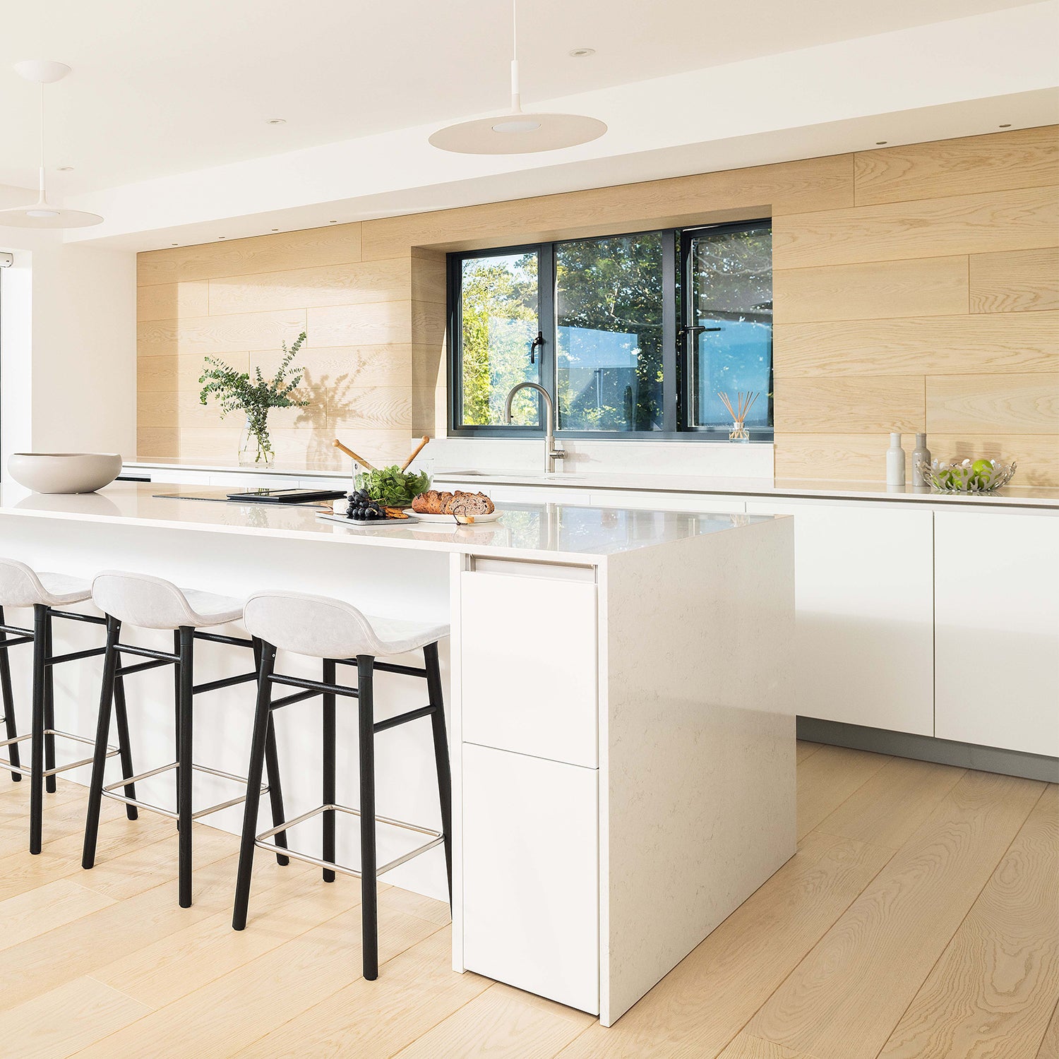 Bright and modern kitchen design by Iroka Interiors featuring sleek white handleless cabinetry, waterfall island with breakfast bar seating, light oak flooring, and minimalist styling – previous bespoke kitchen project in Cornwall.