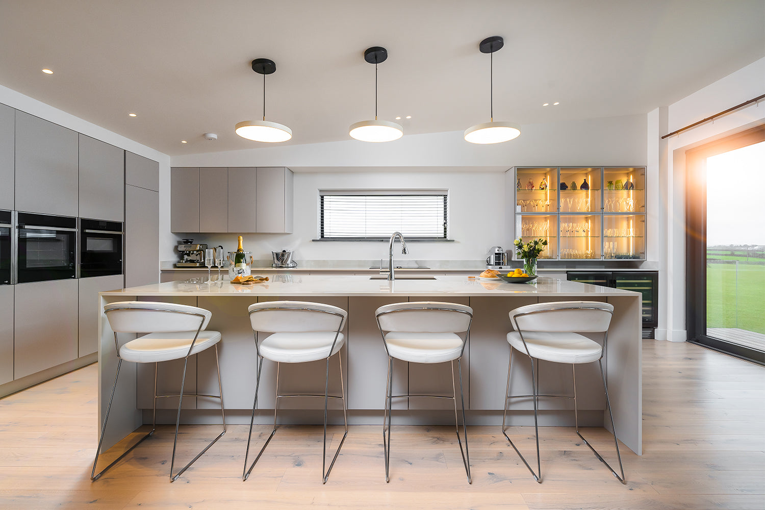 Contemporary open-plan kitchen with soft grey handleless cabinetry, large central island with quartz worktop, integrated appliances, illuminated glassware display, and sleek chrome bar stools – bespoke kitchen design and installation by Iroka Interiors Cornwall.