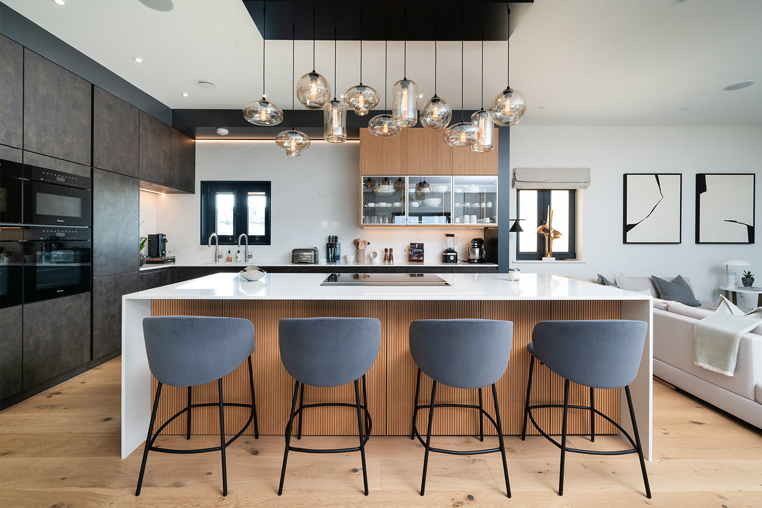 Contemporary open-plan kitchen with statement pendant lighting, sleek white quartz island, integrated appliances, wood panelling, and navy velvet bar stools – bespoke kitchen design and installation by Iroka Interiors Cornwall.