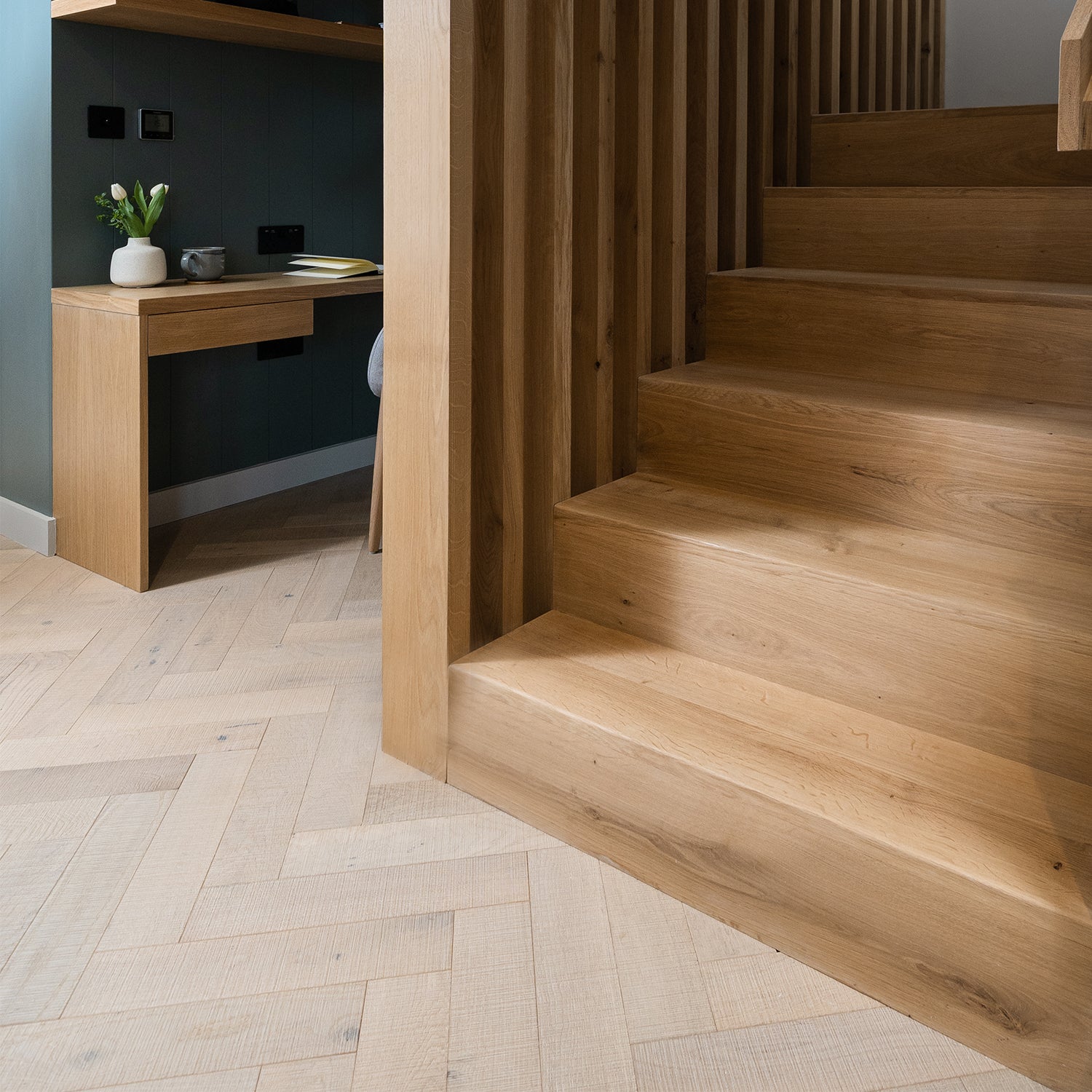 Modern oak staircase with vertical slat balustrade, light herringbone wood flooring, and built-in home office nook featuring minimalist oak desk – contemporary hallway and under-stair design inspiration.