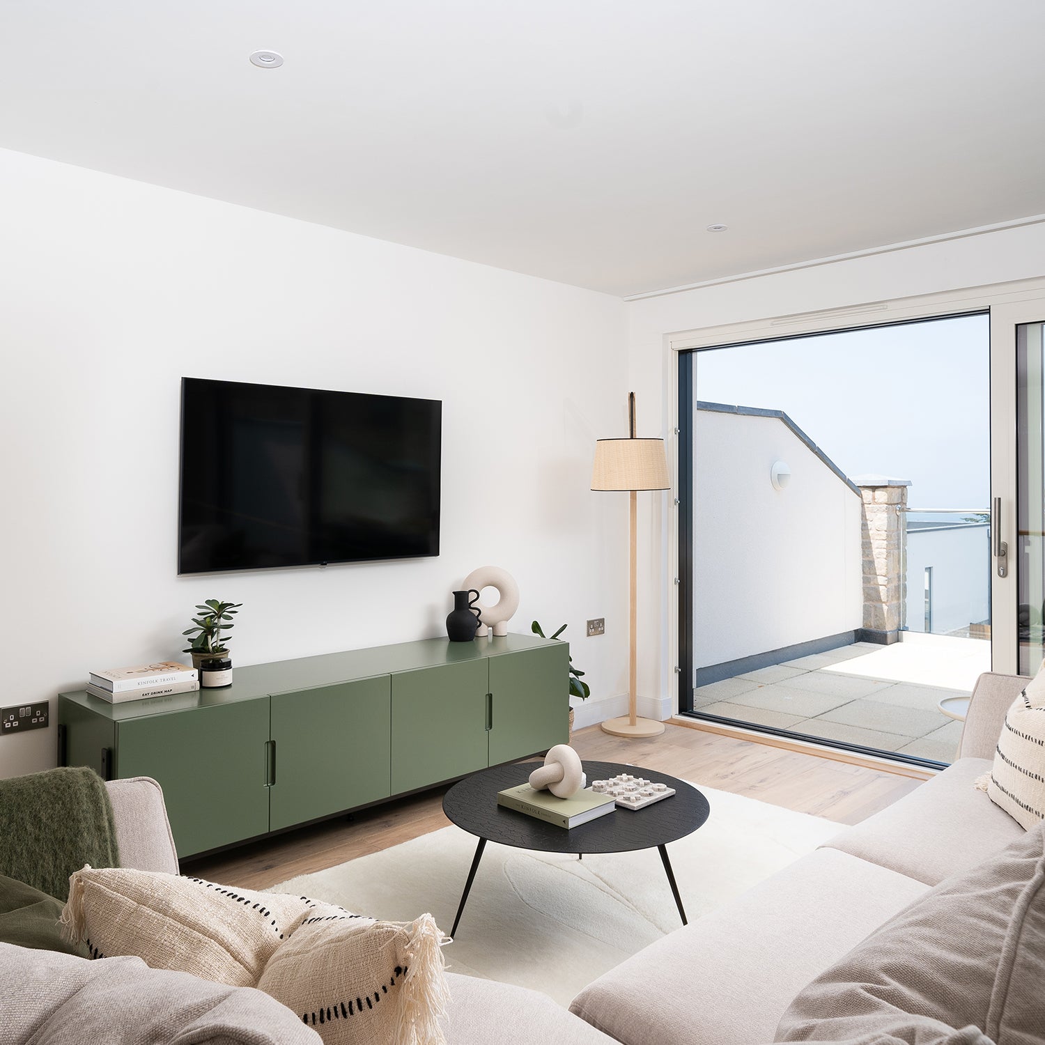 Modern minimalist living room with a neutral-toned sofa, green media console, wall-mounted flat-screen TV, black round coffee table, and sliding glass doors opening to a terrace with sea views.