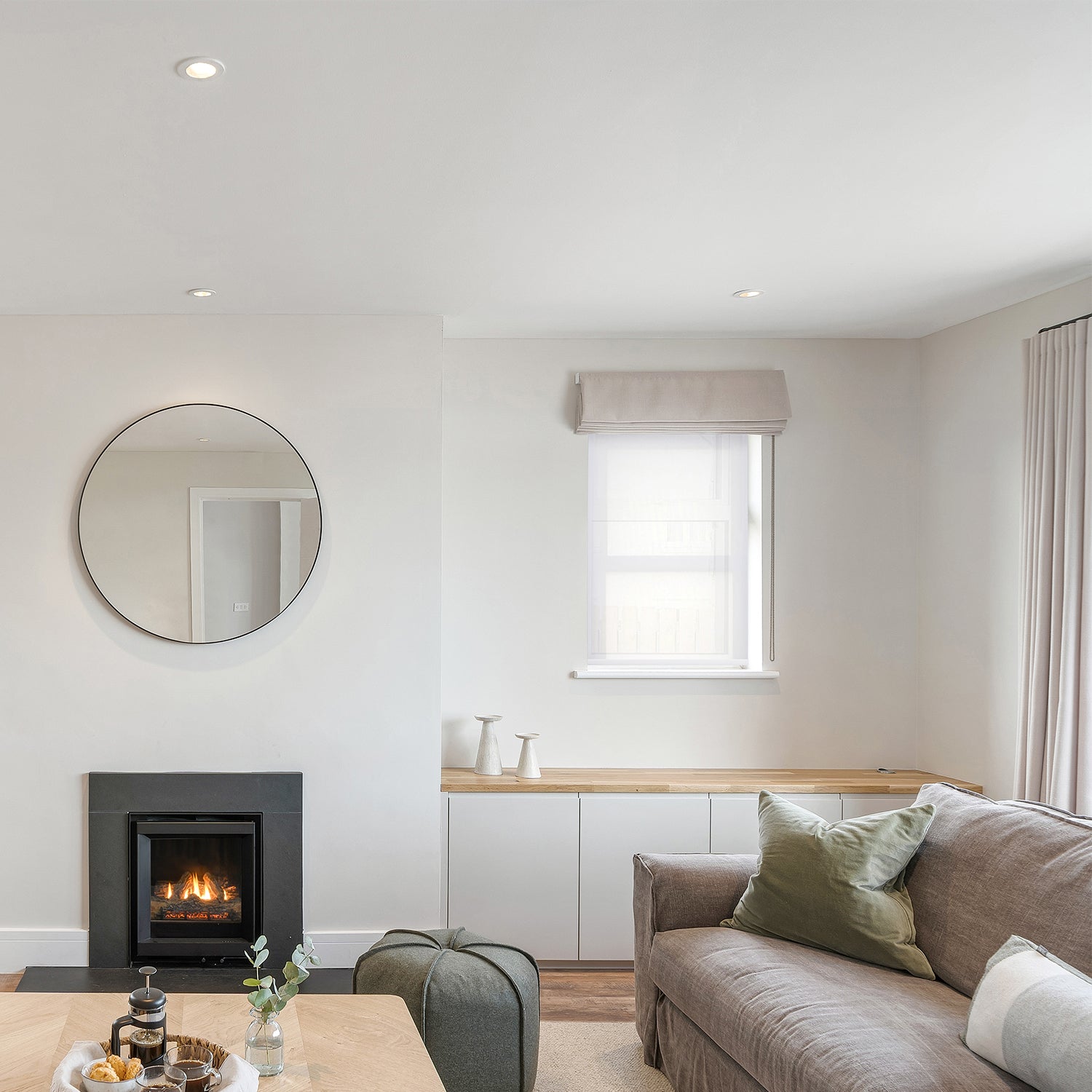 Bright and contemporary living room featuring a neutral colour palette, Roman blind and wave curtains, a modern black wood-burning stove, soft taupe sofa with sage green cushions, and a round wall mirror above a built-in bench.