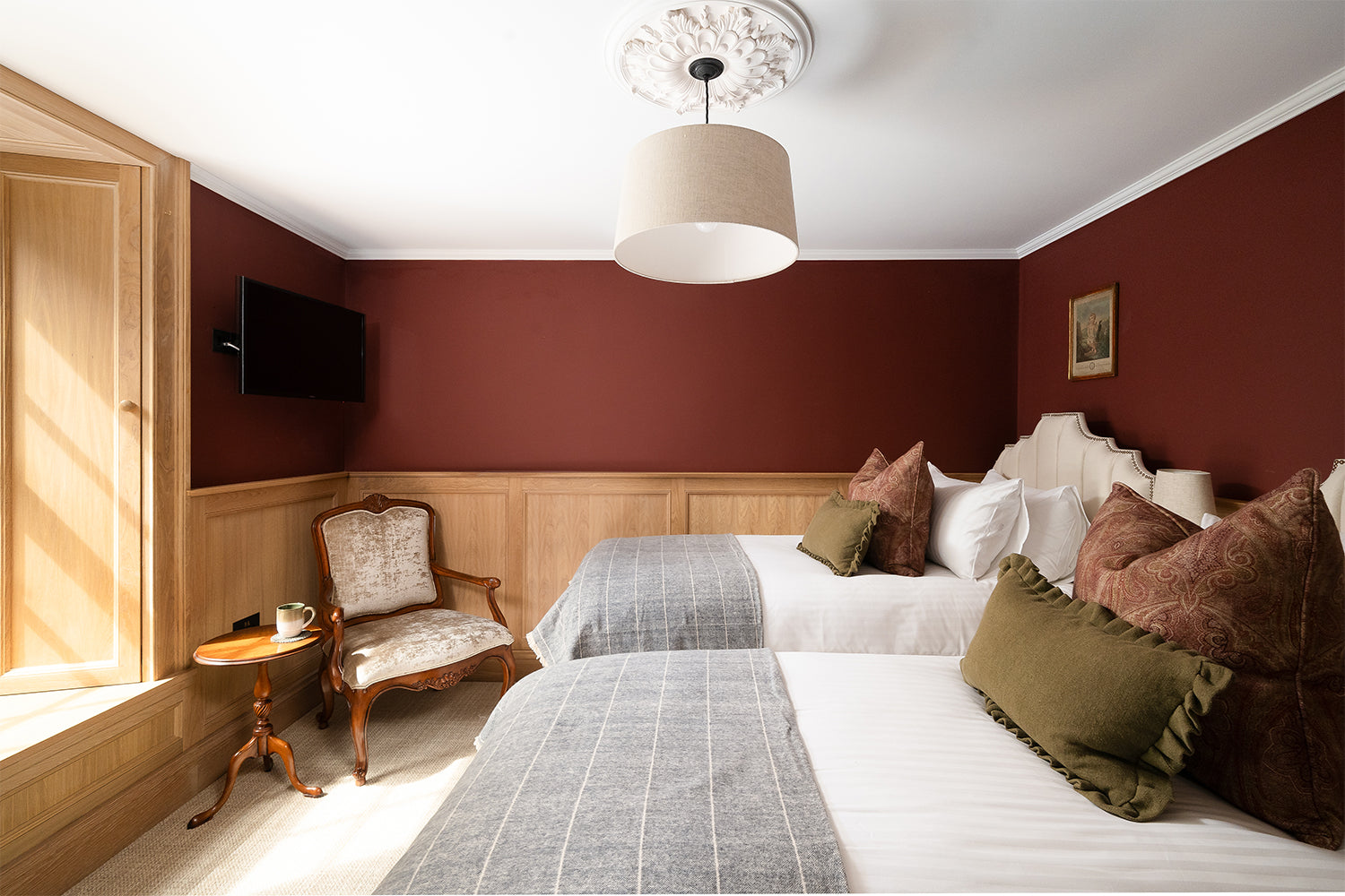 Elegant twin bedroom at Mariners, St Ives featuring upholstered headboards, layered linen bedding in earthy tones, deep burgundy walls with oak panelling, vintage carved wood armchair, and a ceiling rose with modern pendant light - blending period character with boutique hotel comfort.