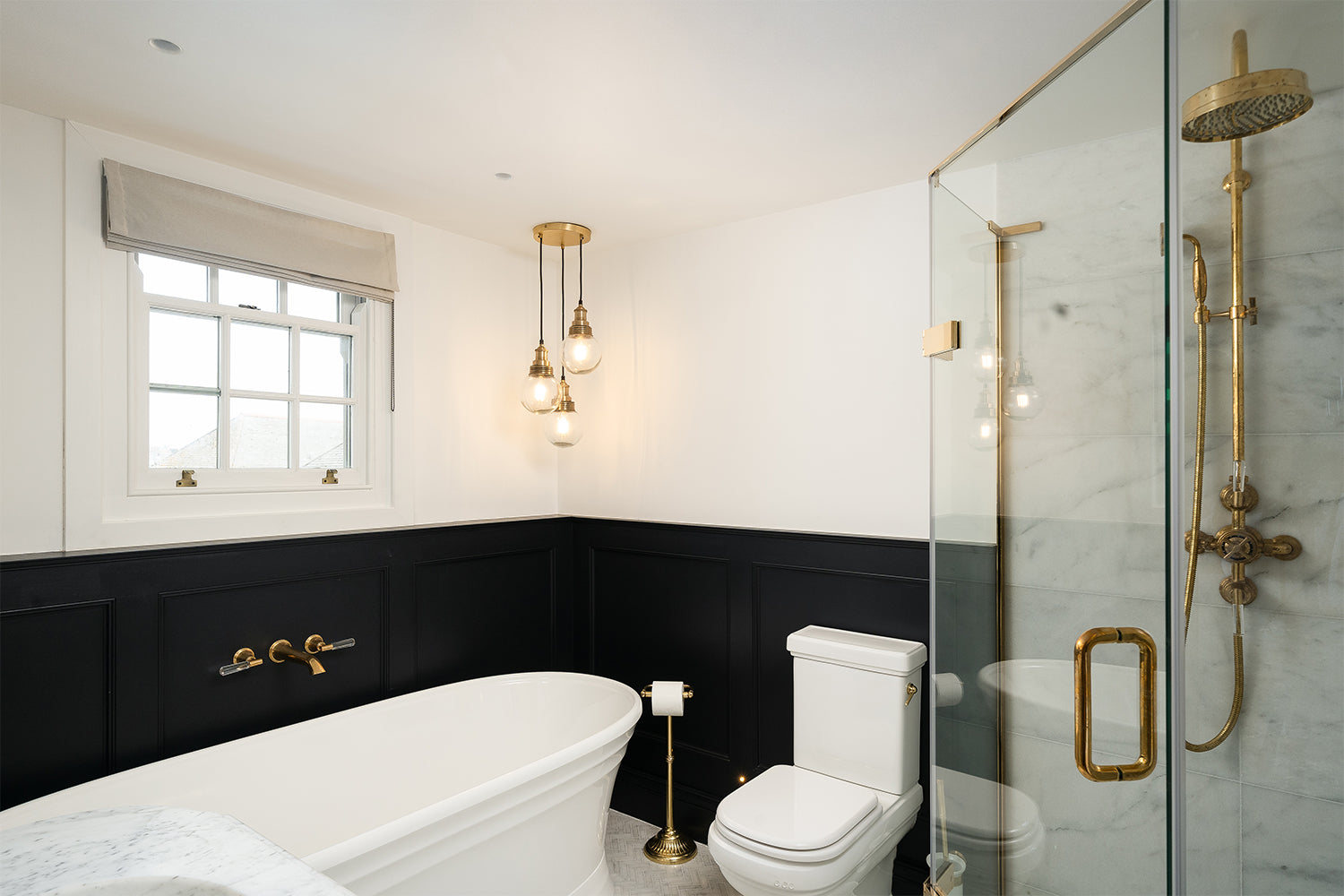 Stylish bathroom at Mariners, St Ives featuring a freestanding roll-top bathtub with wall-mounted brass tapware, black lower wall panelling, pendant bulb lighting, marble walk-in shower with gold fixtures, and traditional white, combining heritage charm with luxury finishes.