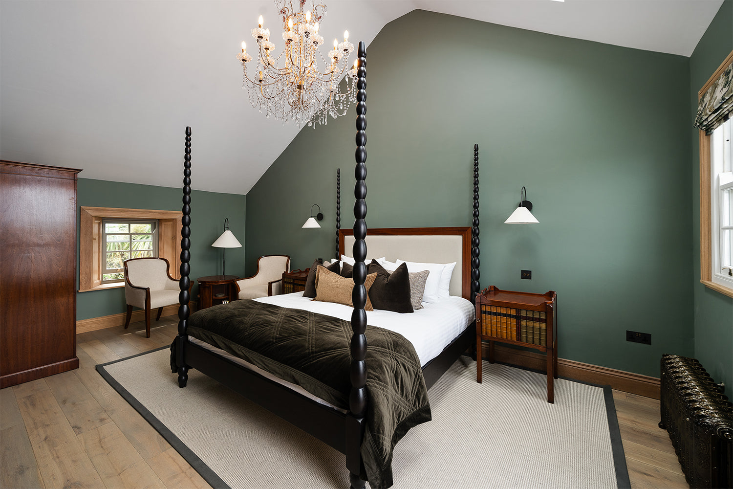 Elegant four-poster bedroom at Mariners, St Ives featuring a statement black spindle bed frame, olive green walls, crystal chandelier, classic wooden furniture, and soft neutral accents—combining period charm with contemporary luxury.