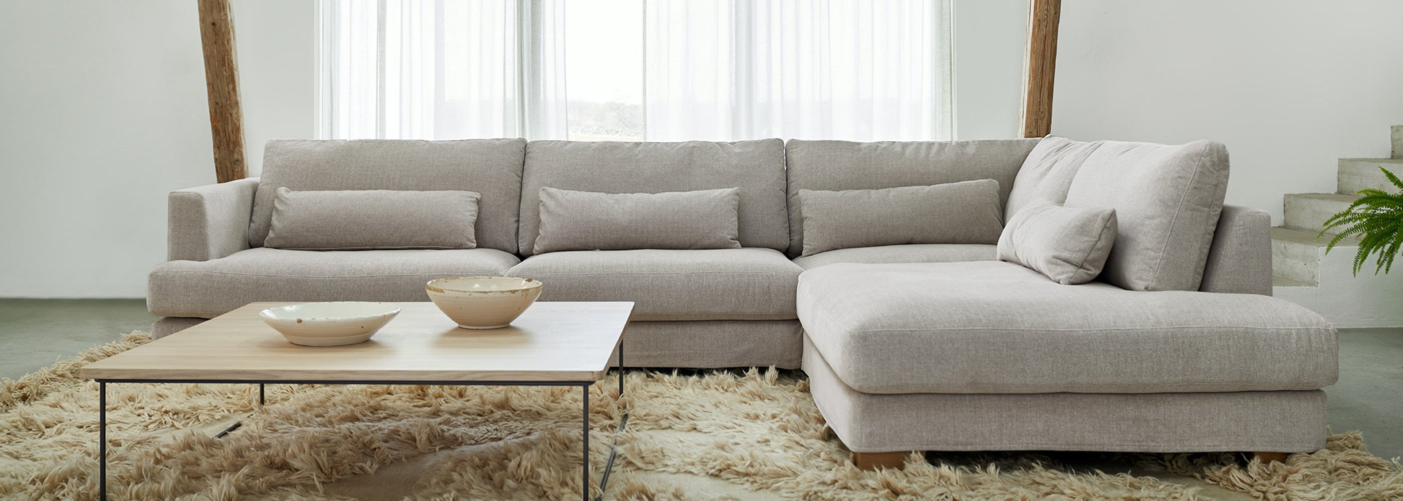Minimalist living room with a light grey Mastrella corner sofa, low wooden coffee table, and ceramic bowls atop a deep shaggy rug, styled with neutral tones and natural textures.