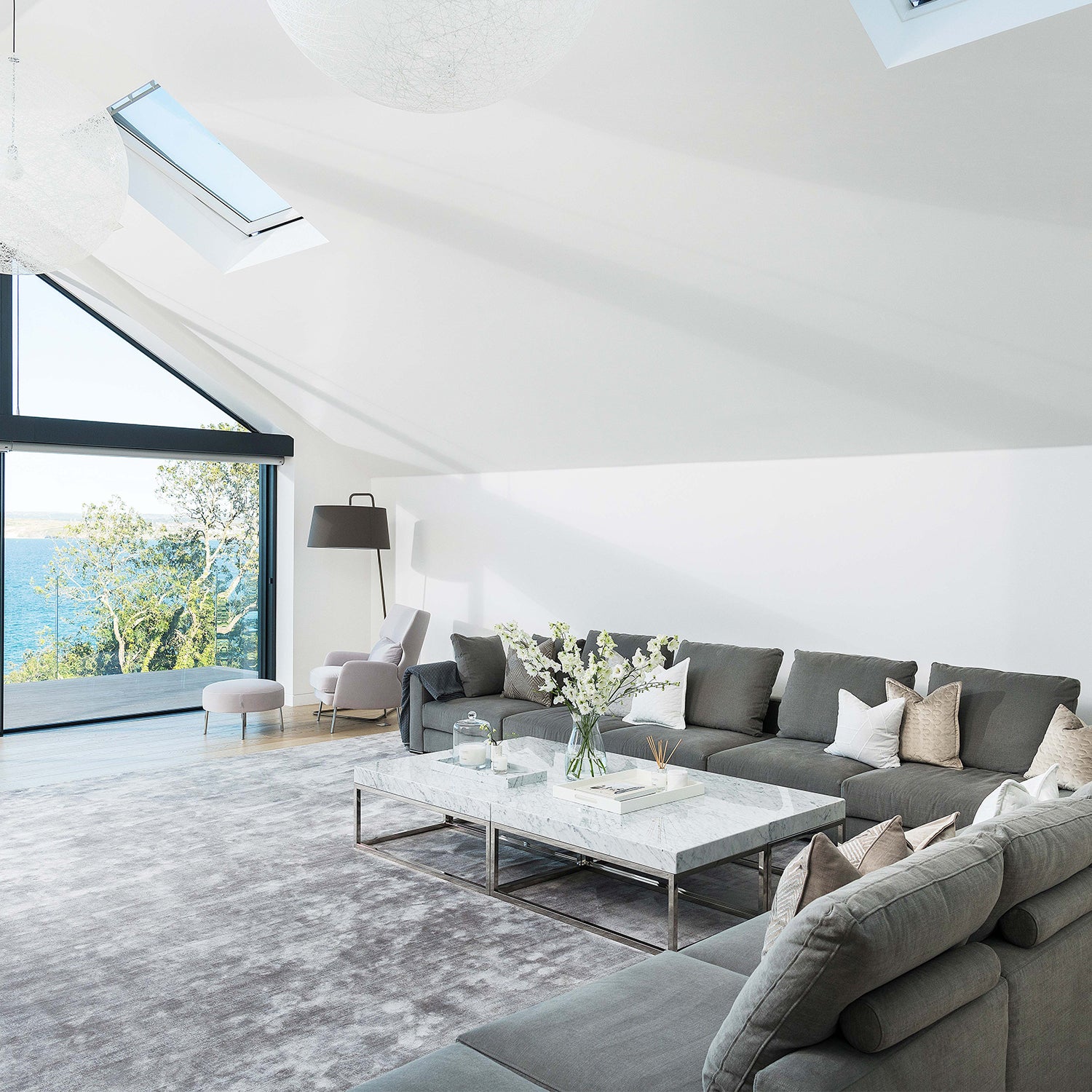 Luxury coastal living room in Cornwall with panoramic sea views, modern grey sectional sofa, marble coffee table, vaulted ceiling with skylights, and soft neutral accents – bespoke interior design and premium furniture by Iroka Interiors.