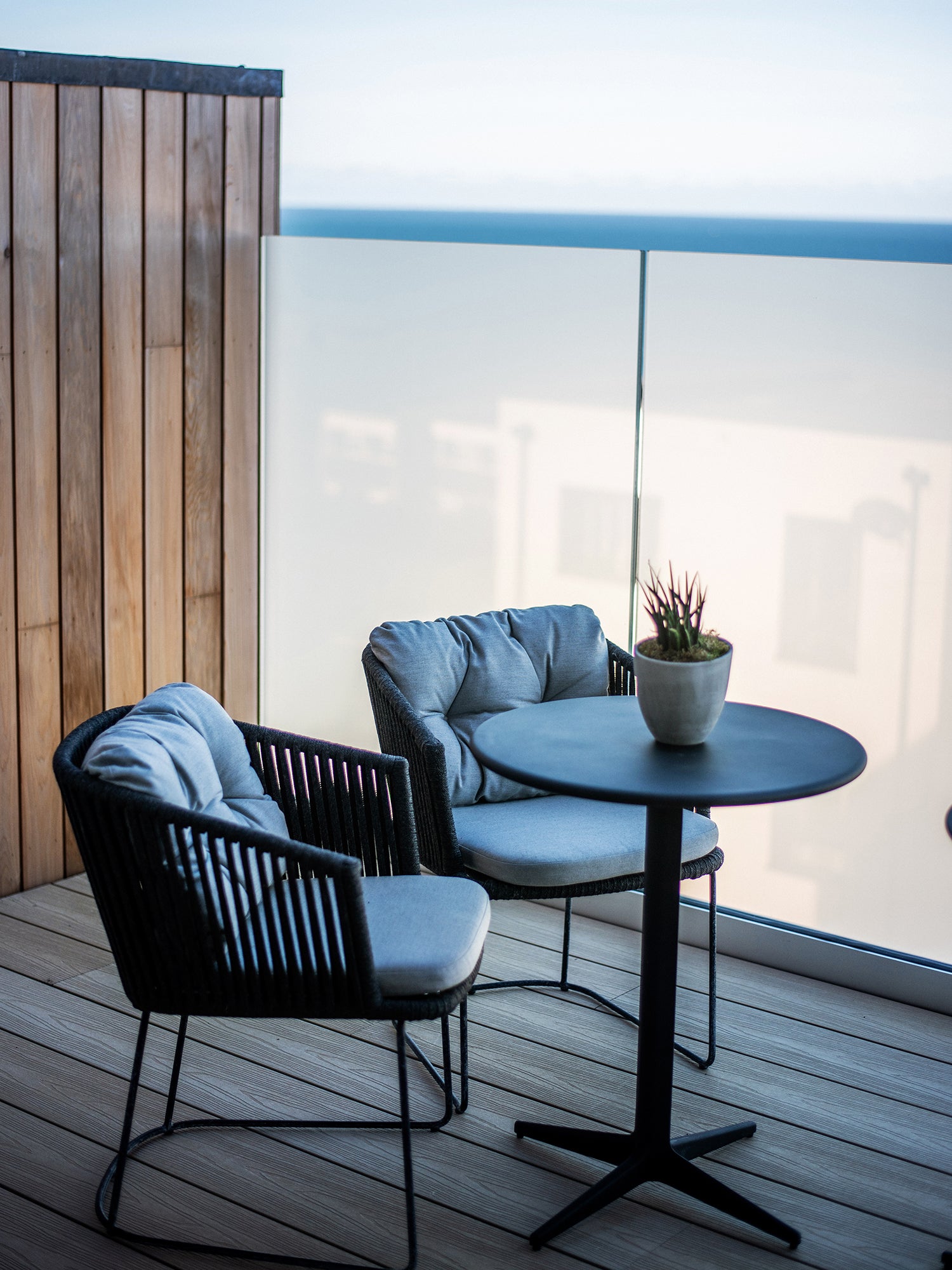 Modern balcony seating area featuring two black-framed lounge chairs with grey cushions, a round black café table, and a potted succulent, set against a frosted glass balustrade with sea views.