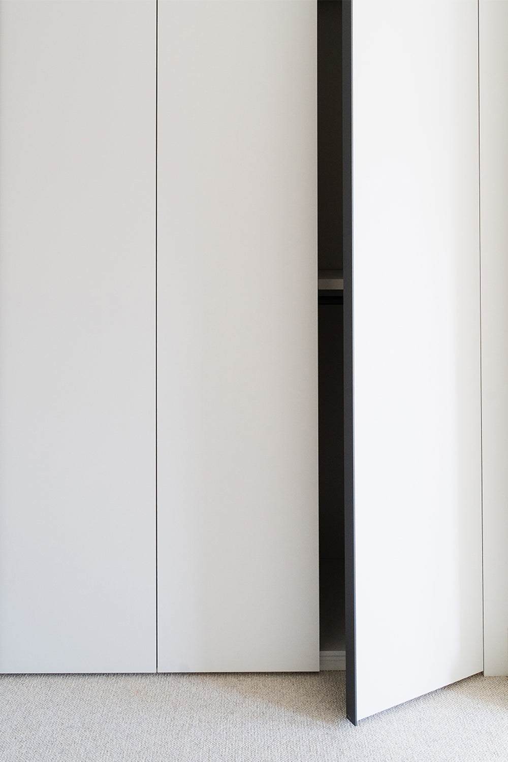 Minimalist built-in white wardrobe with partially open flush doors, showcasing seamless joinery against a neutral carpeted floor.