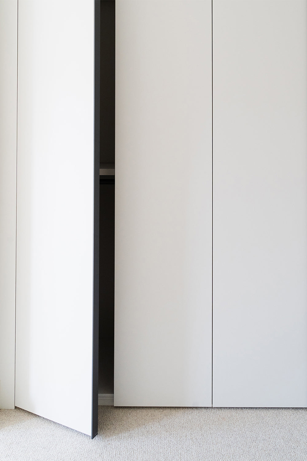 Contemporary white built-in wardrobe with a partially open handleless door, revealing a shadowed interior and minimalist detailing against a light carpeted floor.
