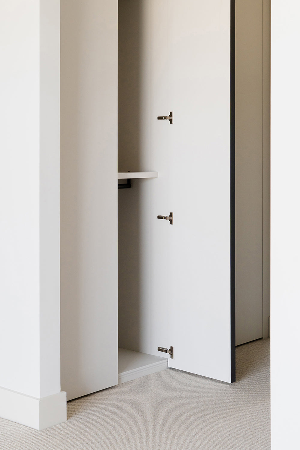 Minimalist white wardrobe with open door, integrated shelf, and concealed hinge hardware, set against a neutral-toned wall and beige carpet flooring.