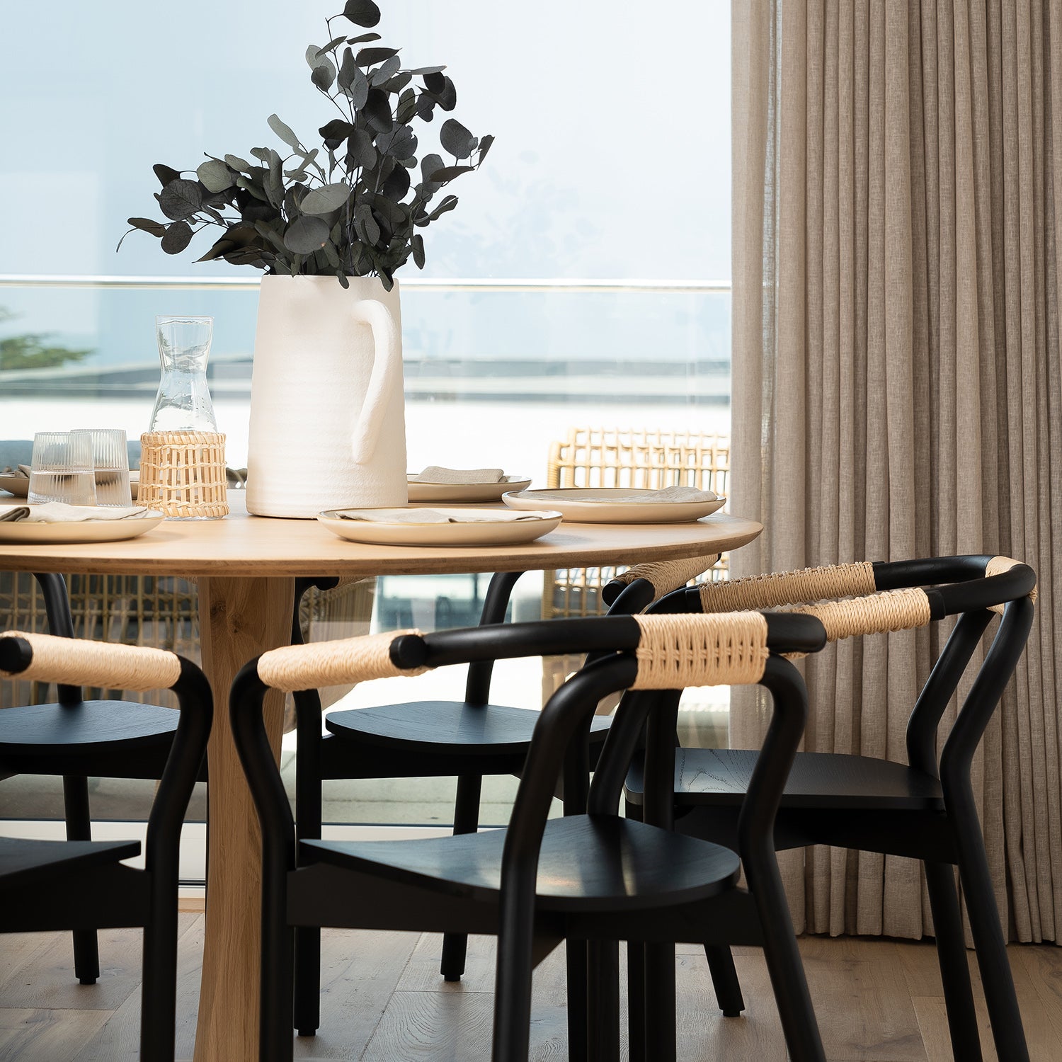 "Modern dining area with natural oak round table, black and rattan dining chairs, neutral linen curtains, and ceramic pitcher centerpiece – contemporary coastal interior design with minimalist Scandinavian styling.
