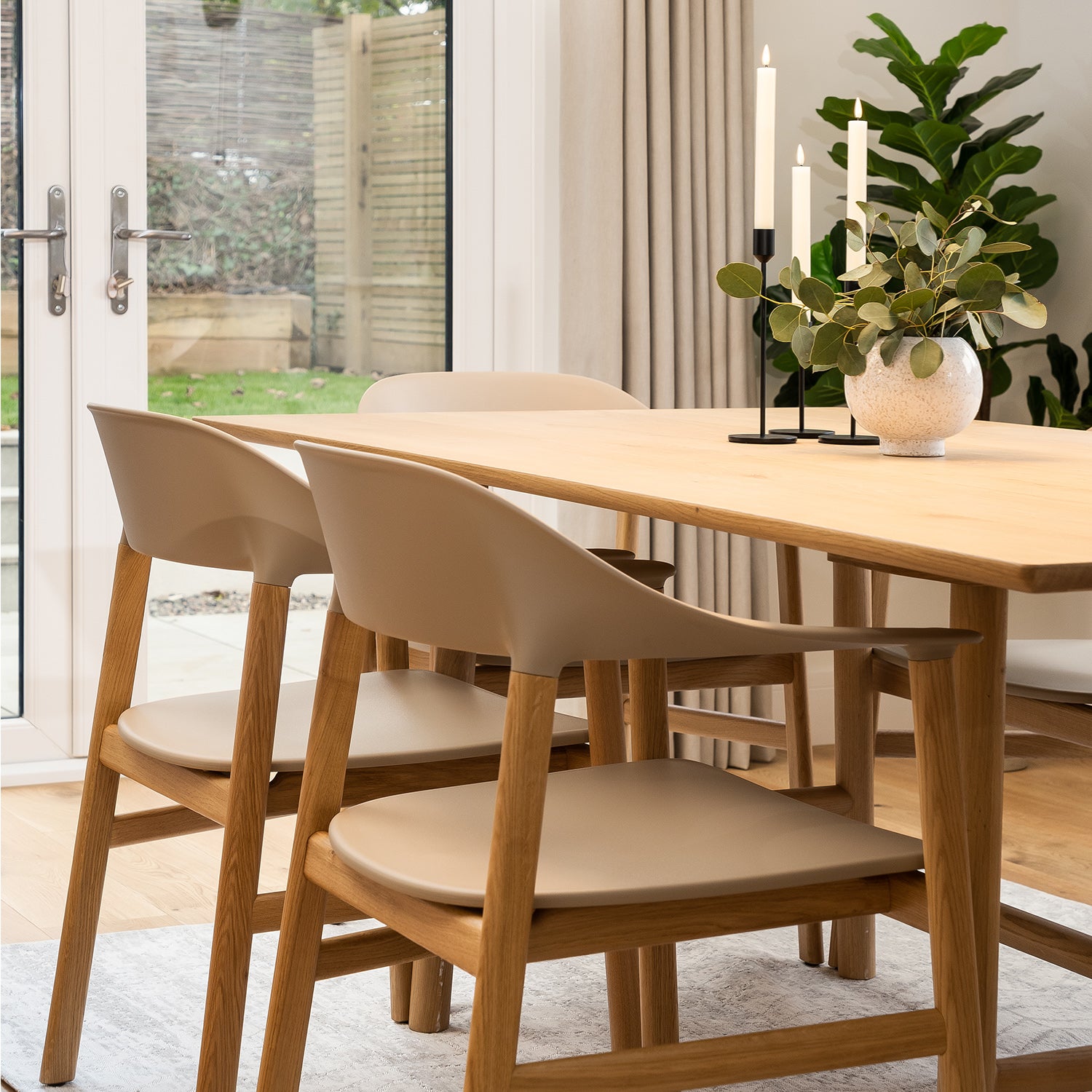 Scandinavian-inspired dining room featuring oak dining table with taupe modern chairs, minimalist black candle holders, neutral curtains, and indoor greenery – contemporary furniture and natural interior styling.