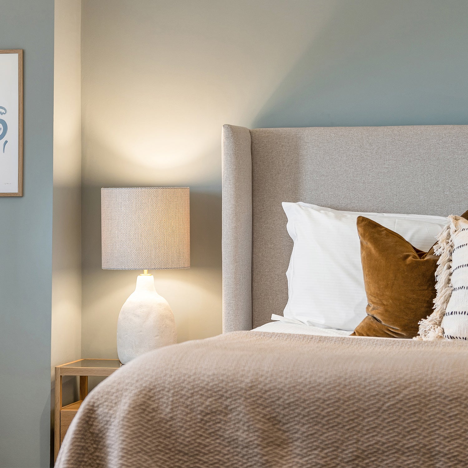 Modern bedroom with light grey upholstered headboard, soft taupe bedding, velvet accent pillow, ceramic table lamp, and muted green wall – cozy and elegant interior design with warm neutral tones.