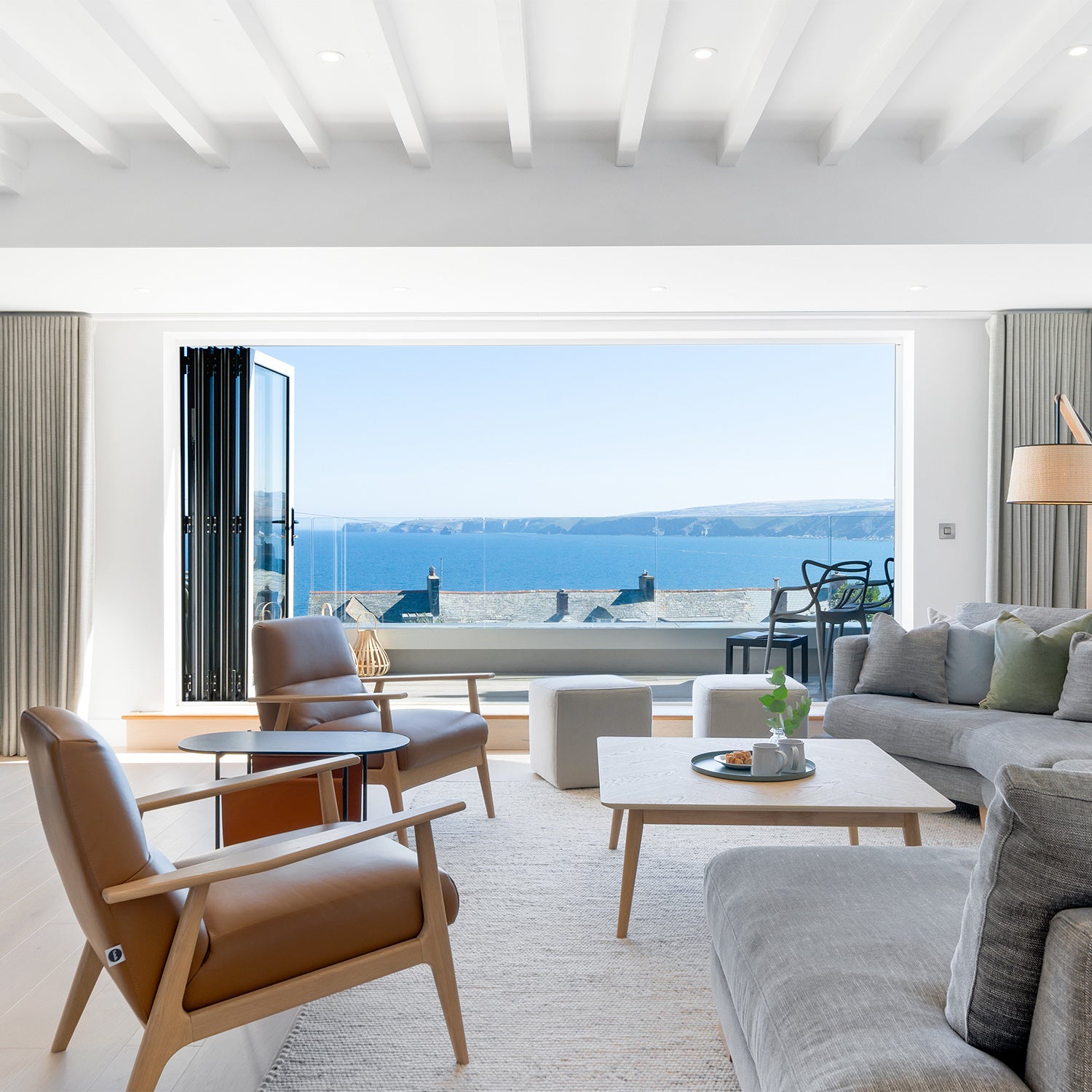 Modern coastal living room with ocean view, featuring grey linen sofas, leather accent chairs, minimalist wood coffee table, and full-width bi-fold doors – contemporary interior design and premium furniture for luxury seaside homes.