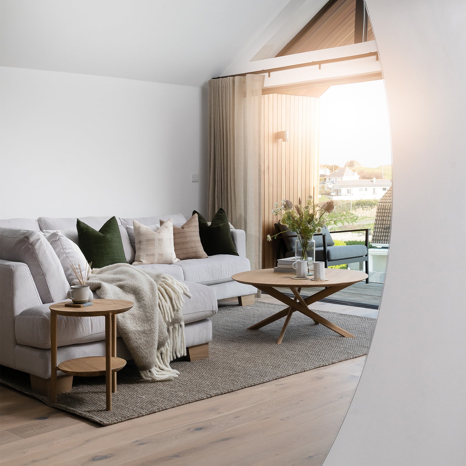 Bright coastal-inspired living room in Cornwall with neutral sofa, layered cushions, wooden armchair, rustic coffee table, and soft natural textures – modern interior design and handcrafted furniture for relaxed seaside homes.