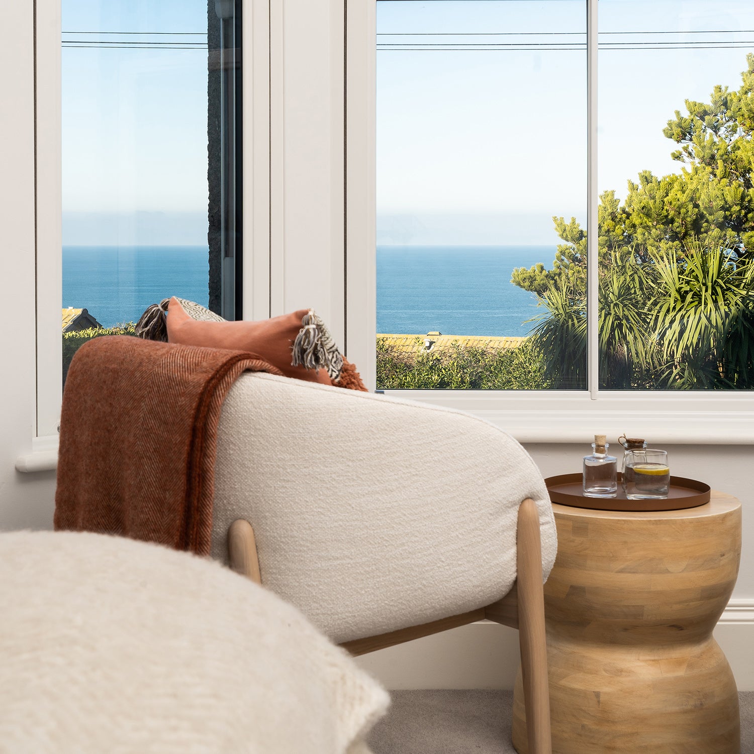 Coastal reading nook with boucle lounge chair, burnt orange throw, wooden side table, and ocean view through large white-framed windows – serene modern interior design with natural textures and calming color palette.