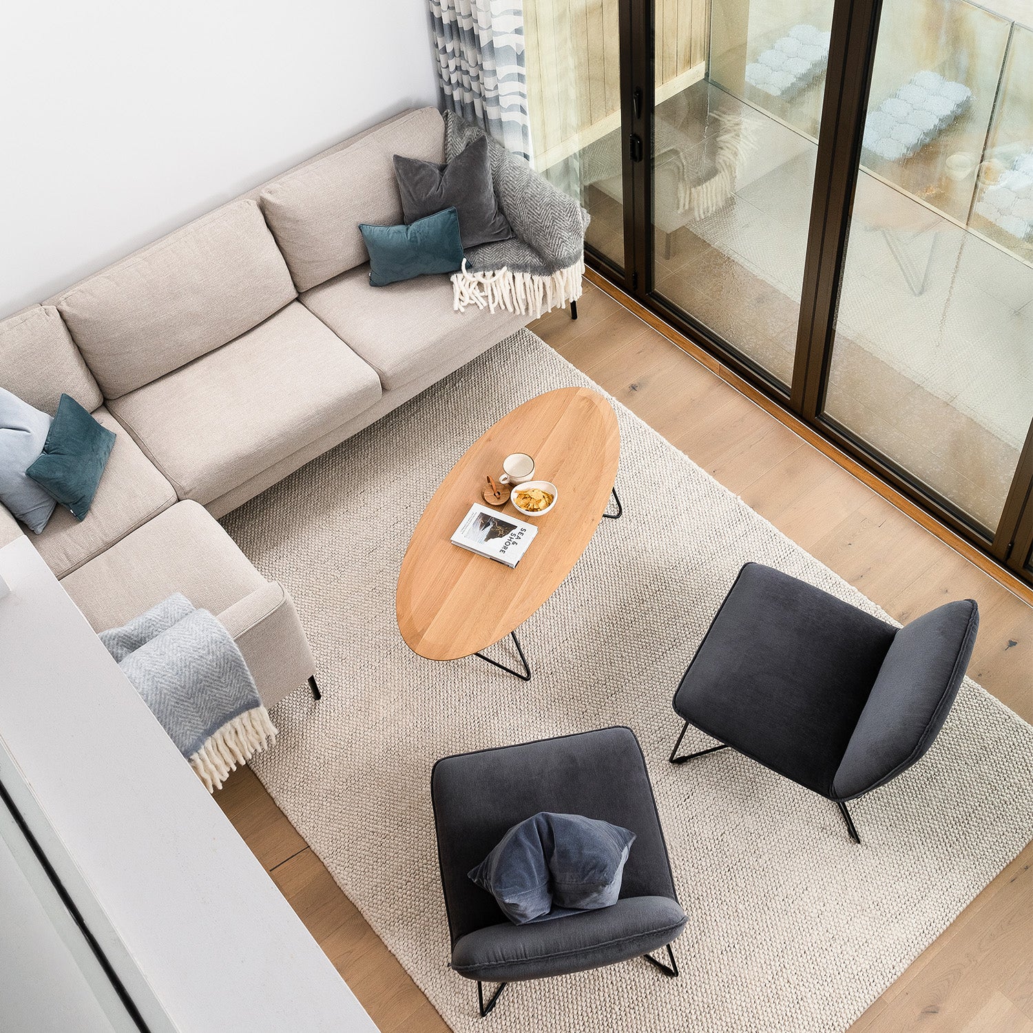 "Modern Cornwall living room with neutral corner sofa, charcoal accent chairs, oval wood coffee table, textured rug, and full-height sliding glass doors – contemporary coastal interior design and bespoke furniture.