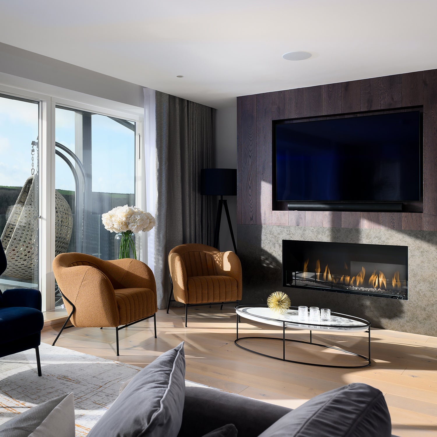 "Contemporary living room with modern gas fireplace, wall-mounted TV, rust-colored accent chairs, marble coffee table, and floor-to-ceiling curtains – sleek interior design with cozy luxury and panoramic patio views.