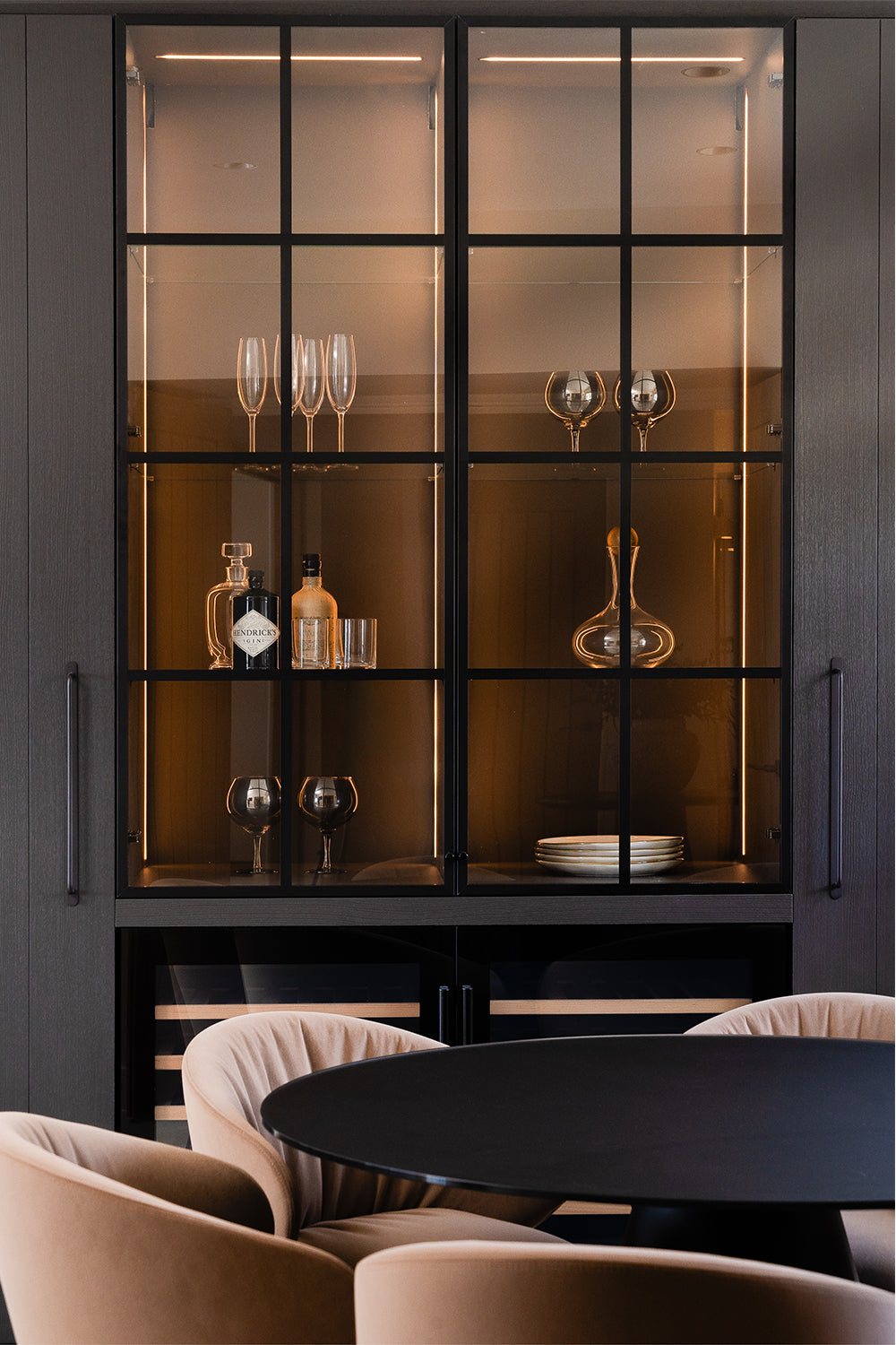 Elegant built-in glass cabinet with interior lighting, showcasing glassware, spirits, and tableware; in front, a round black dining table with soft beige velvet chairs.