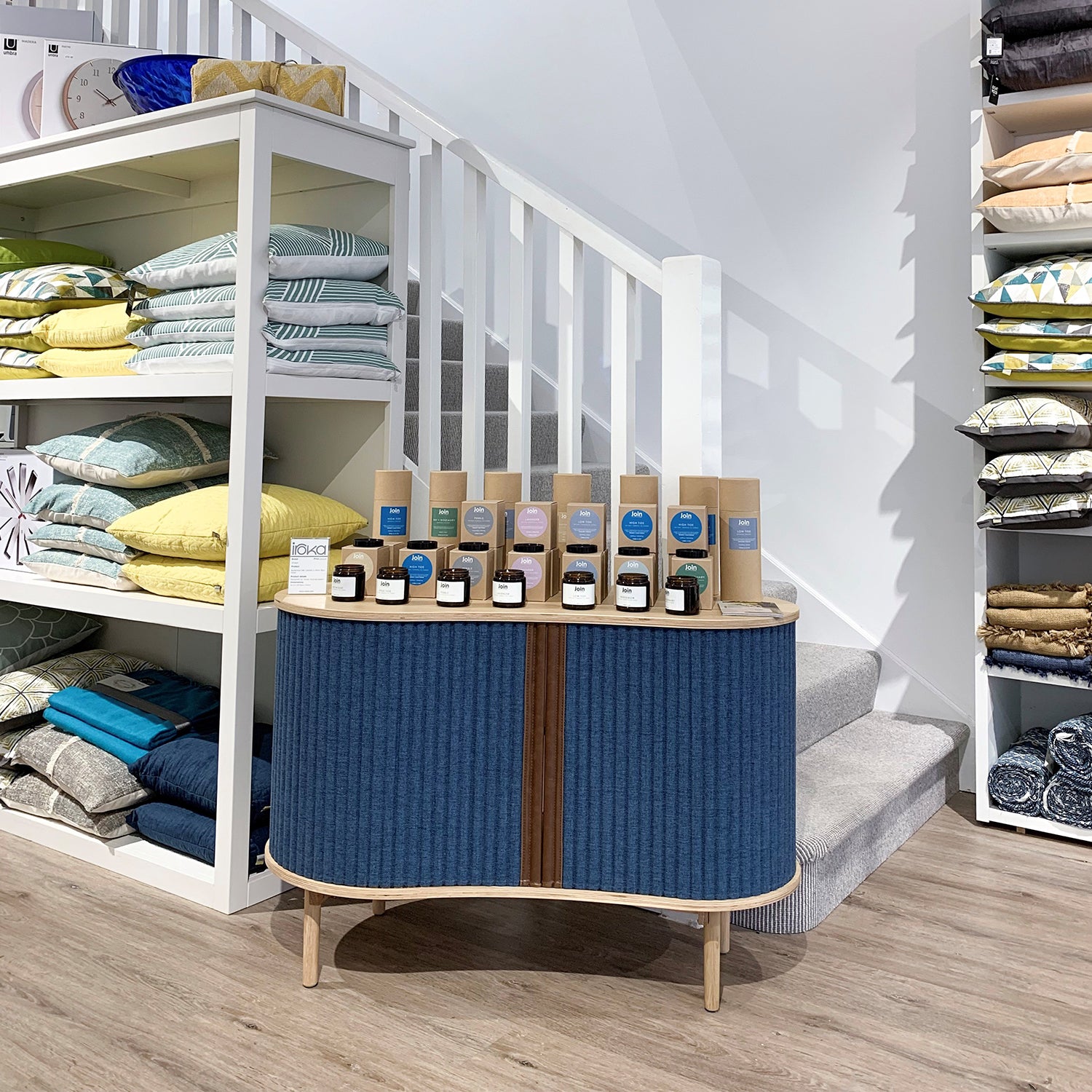 Interior of Iroka Interiors' Wadebridge shop featuring neatly stacked designer cushions, stylish textiles, and curated home fragrance products displayed on a modern blue-fronted console against a bright, contemporary staircase backdrop.
