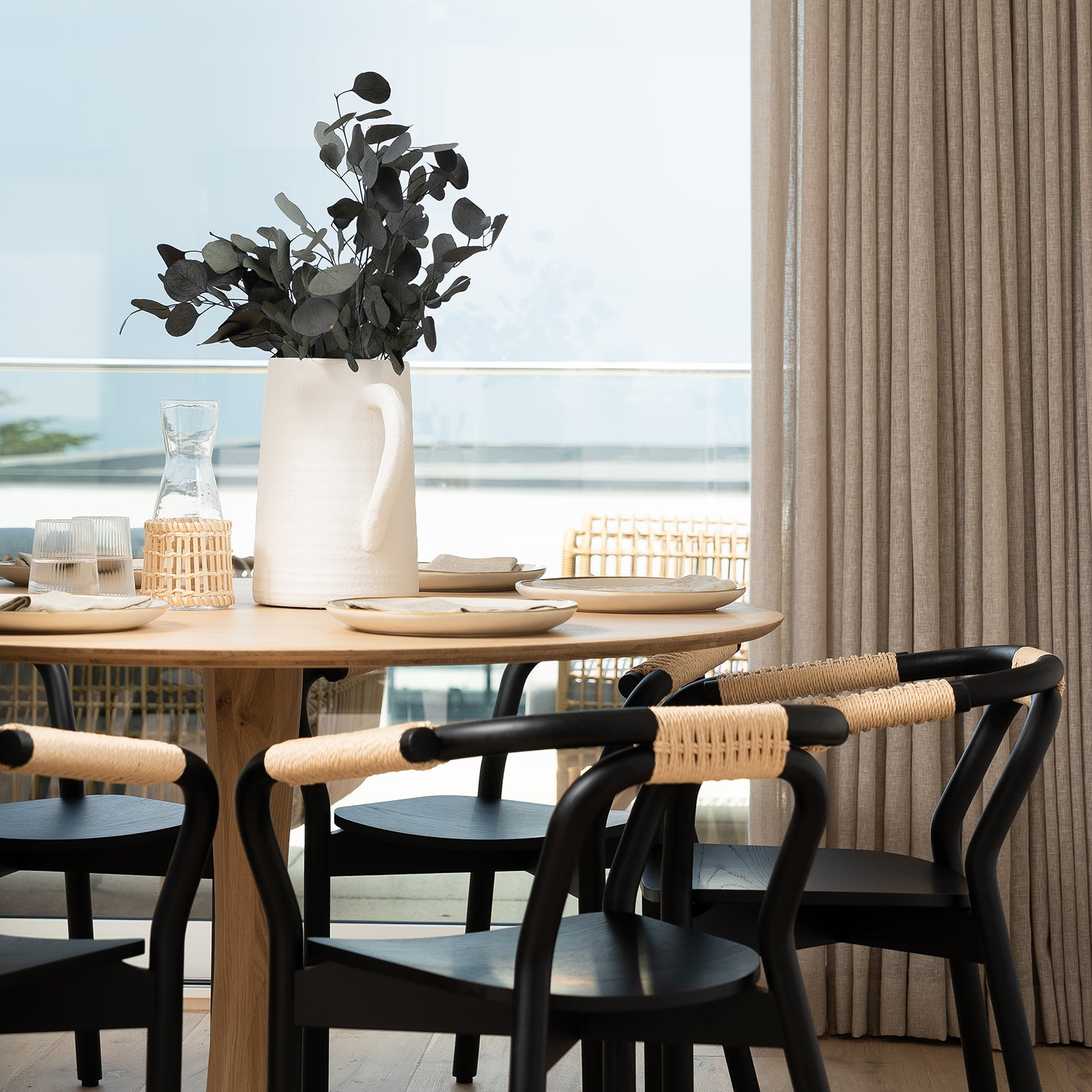 Modern coastal dining area designed by Iroka Interiors in Cornwall, featuring a natural oak round dining table, black wishbone-style chairs with woven backs, and panoramic sea views — blending Scandinavian simplicity with coastal elegance for luxury home interiors.
