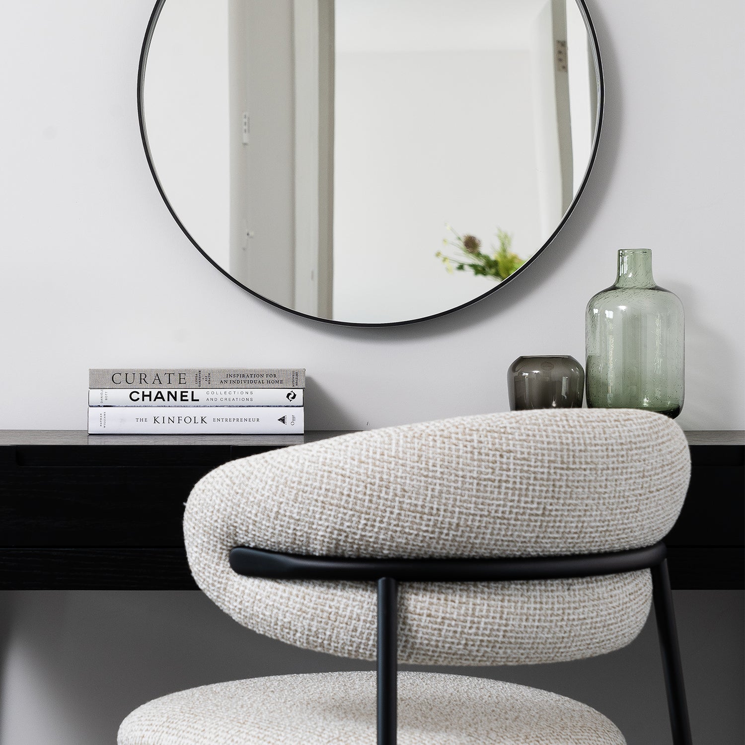 Minimalist home office setup styled by Iroka Interiors in Hayle, Cornwall, featuring a boucle-textured modern chair, black wooden desk, round wall mirror, curated decorative books, and glass vase accents – perfect blend of contemporary furniture and interior design.