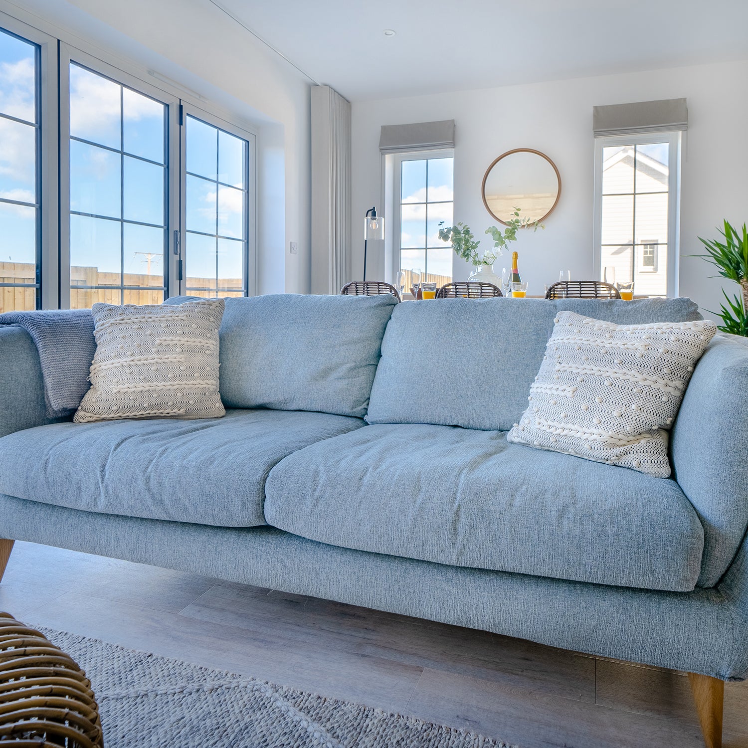 Bright coastal living room in Padstow featuring a powder blue sofa, textured neutral cushions, light wood flooring, and large sash windows – bespoke interior design and handcrafted furniture for Cornish seaside homes.