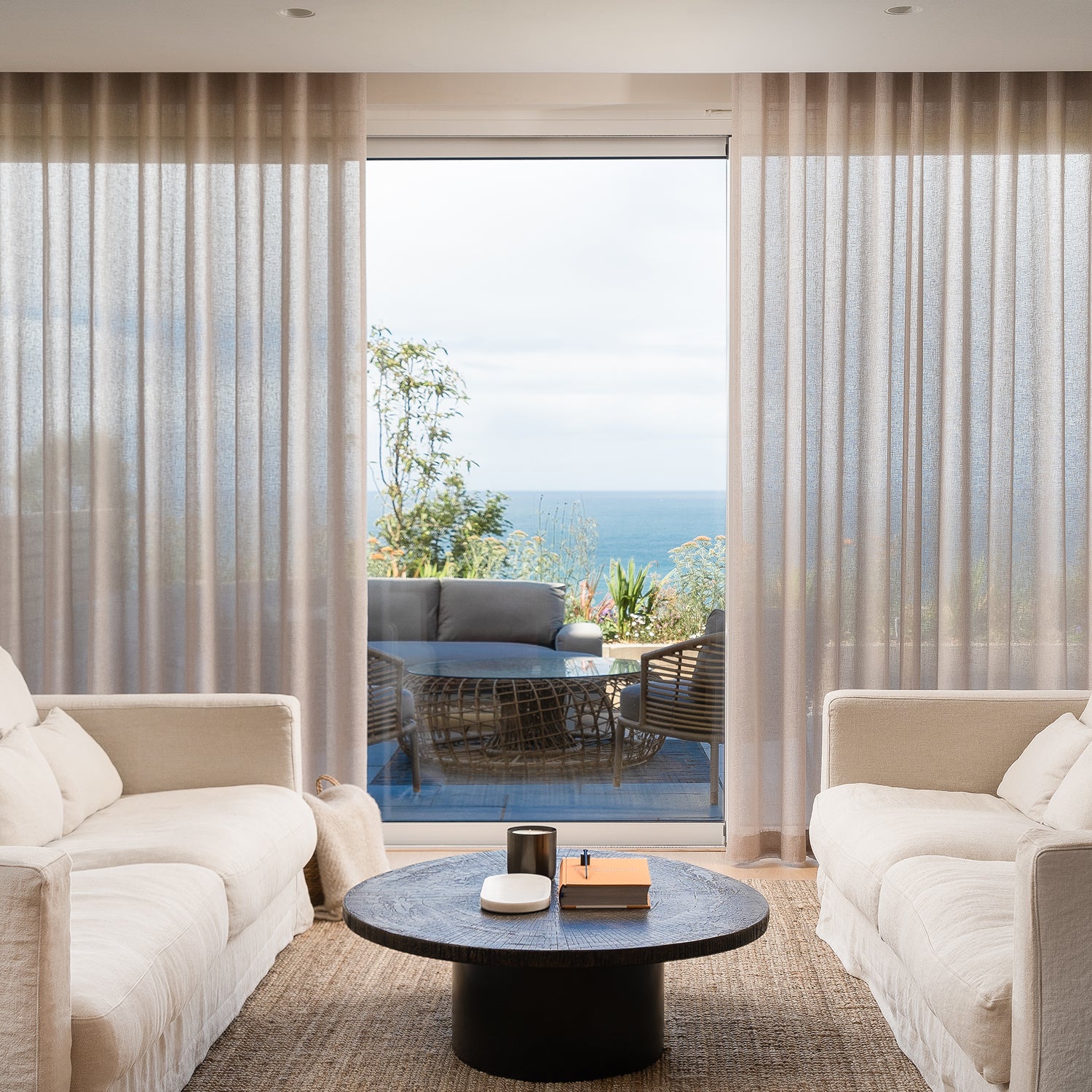 Cornish coastal living room with sheer floor-to-ceiling curtains, cream linen sofas, woven jute rug, round black coffee table, and sliding glass doors opening to ocean-view terrace – modern seaside interior design with natural textures.