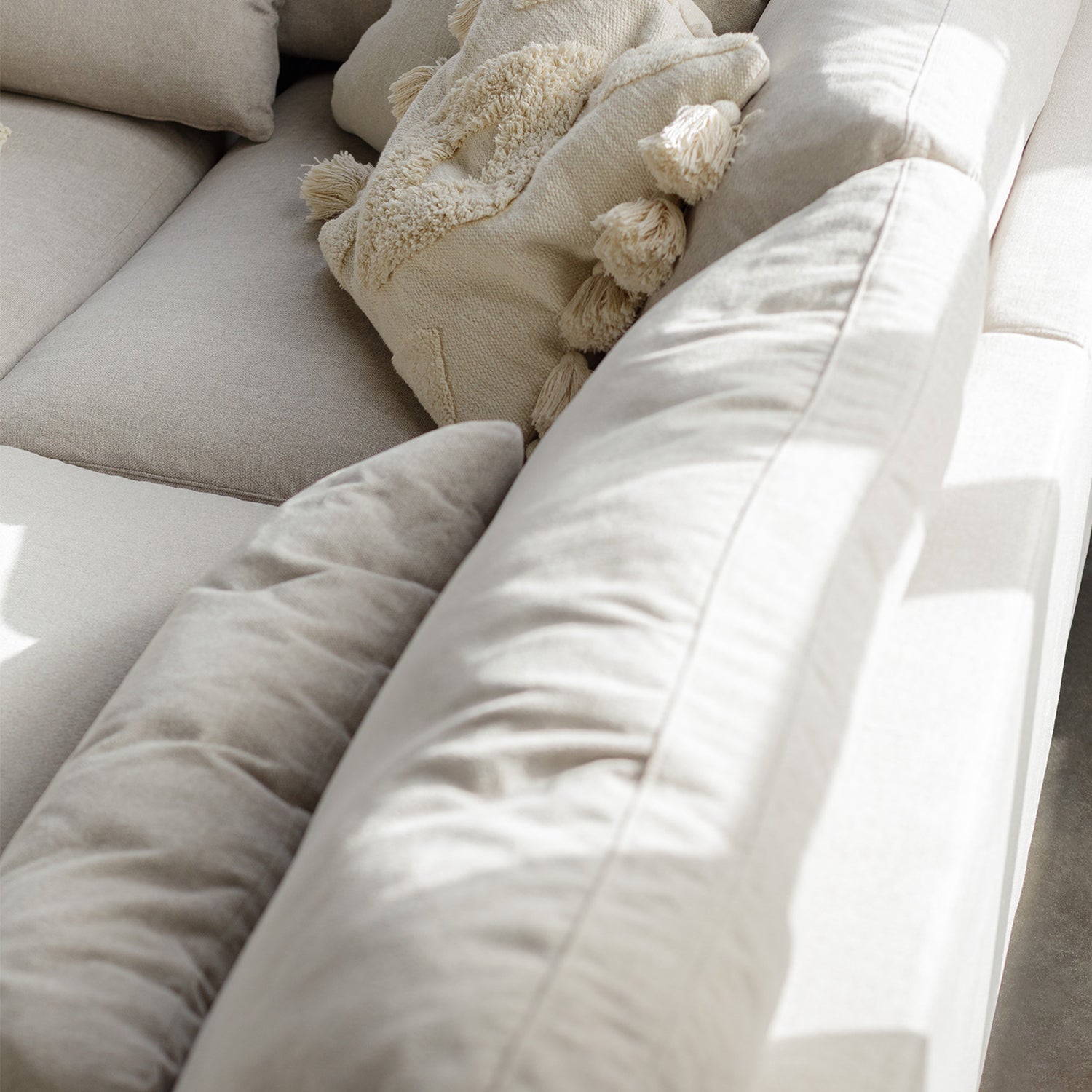 Close-up of soft beige linen sectional sofa with plush back cushions and a textured tassel pillow, bathed in natural sunlight.