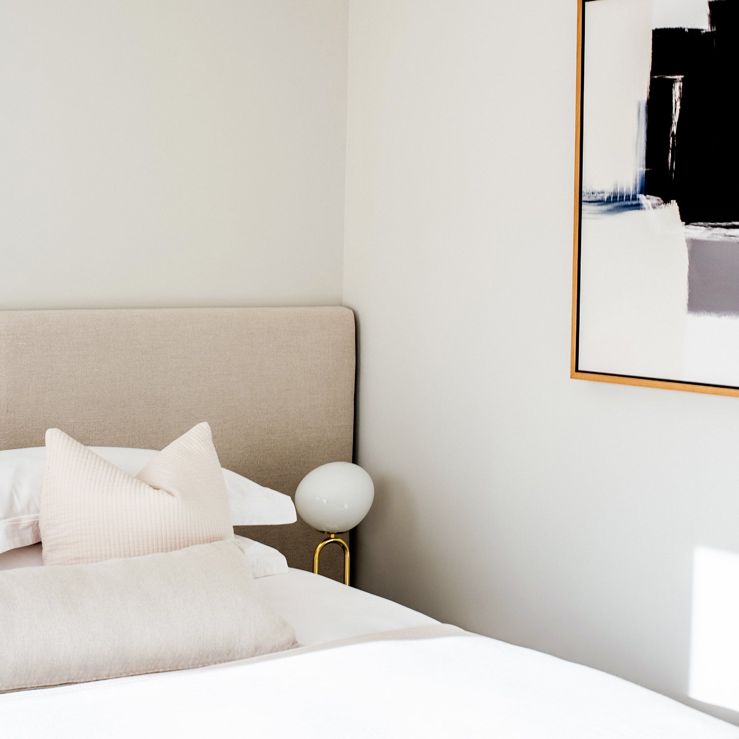 Minimalist bedroom at Pentire Townhouse in Newquay featuring a neutral upholstered headboard, soft layered bedding, contemporary abstract wall art, and a modern orb bedside lamp with brass stand.