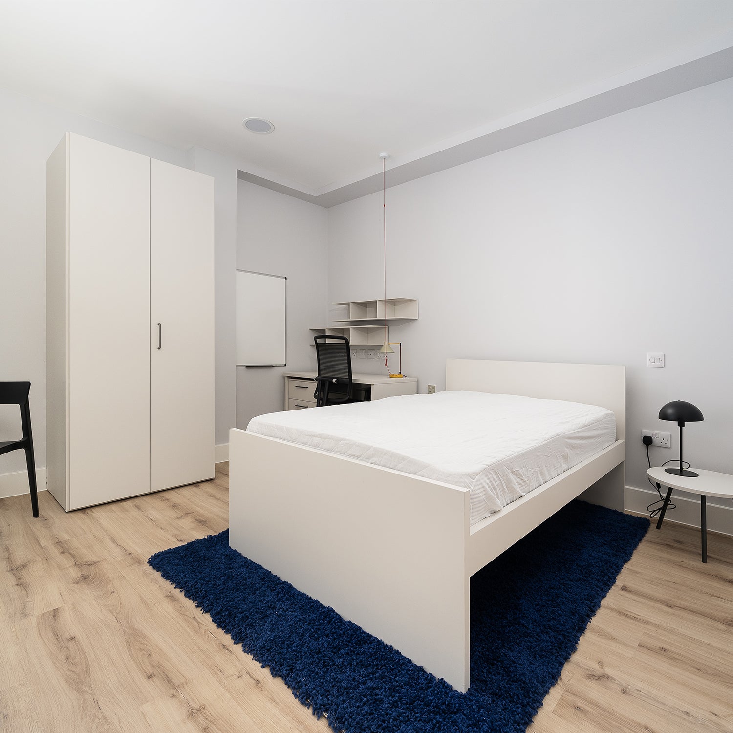 "Stylish and functional student bedroom design in Falmouth by Iroka Interiors, featuring minimalist white furniture, dedicated study space with ergonomic chair and shelving, and a bold blue rug adding contrast and comfort.