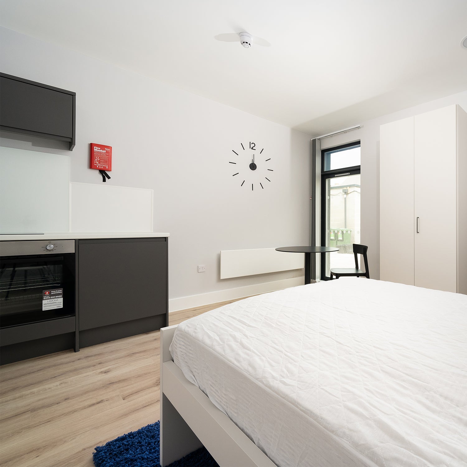Modern Falmouth student studio interior by Iroka Interiors, showcasing a multifunctional layout with integrated kitchen units, sleek black dining area, built-in wardrobe, and contemporary wall clock for functional, space-saving student living.