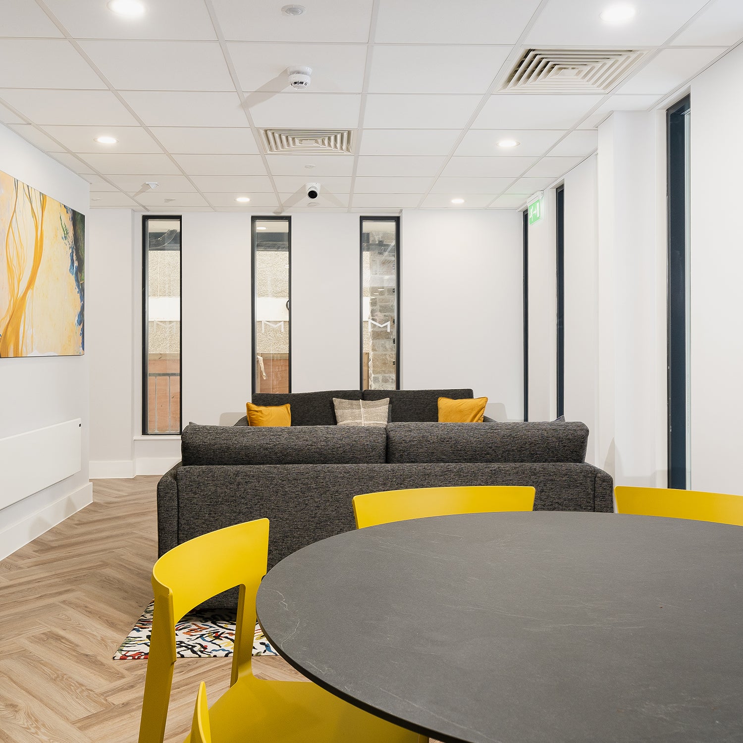 Modern communal lounge and dining area in Falmouth student accommodation by Iroka Interiors, featuring charcoal grey sofas, bold yellow chairs, minimalist decor, and natural light for stylish and functional shared living spaces.
