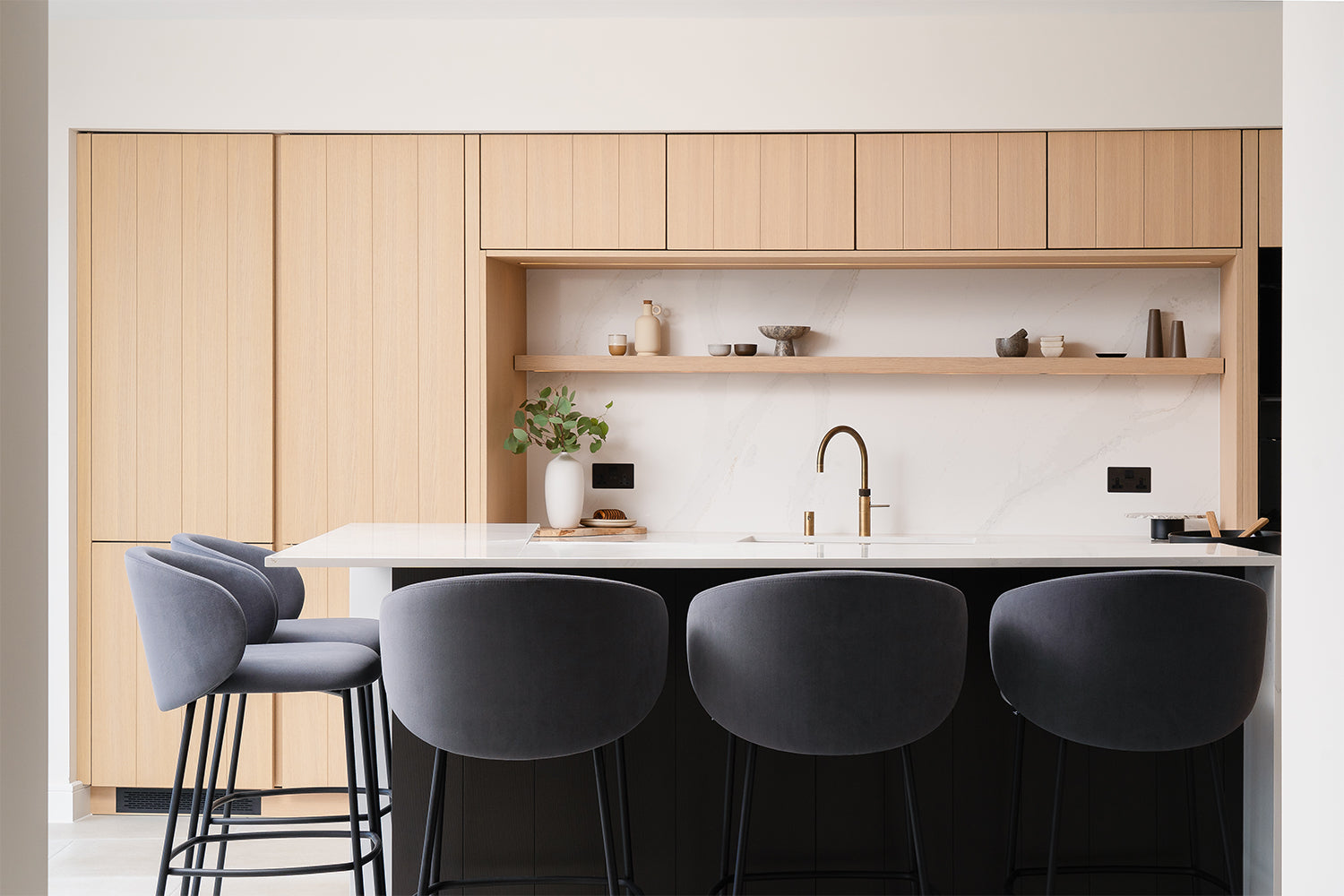Designer kitchen with light wood cabinetry, white quartz island, black panel base, and navy velvet bar stools – sleek contemporary kitchen design with minimalist open shelving and brass fixtures.