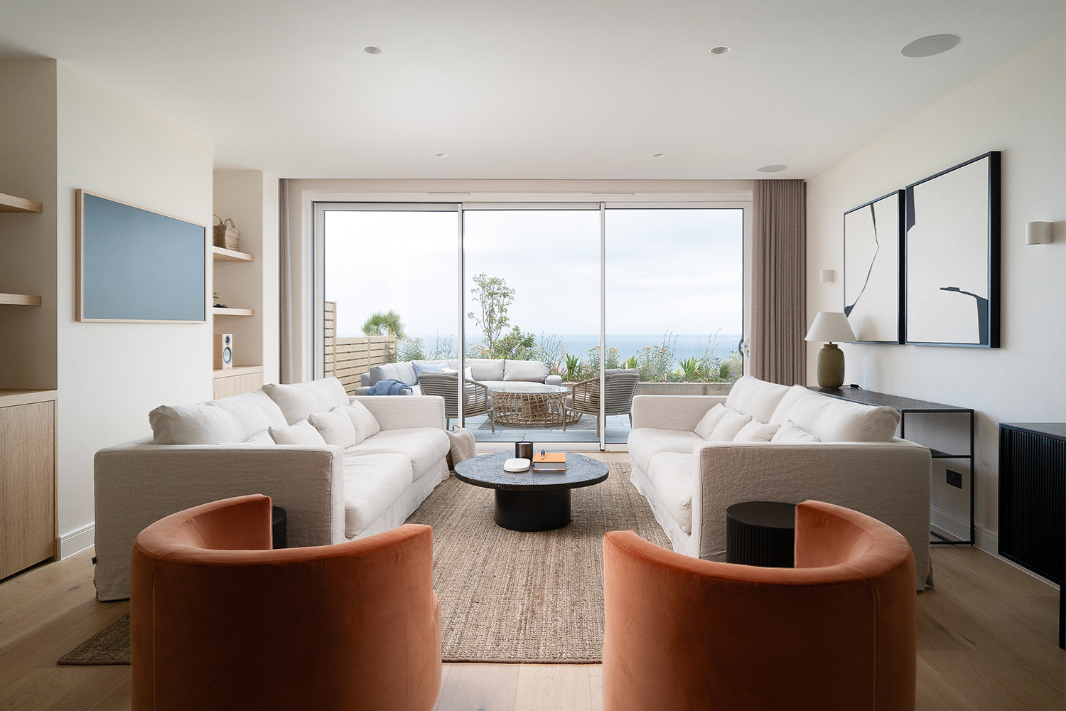 Modern coastal living room with white linen sofas, terracotta velvet armchairs, abstract wall art, sliding glass doors, and ocean-view terrace – cornish seaside interior design with neutral tones and natural textures.