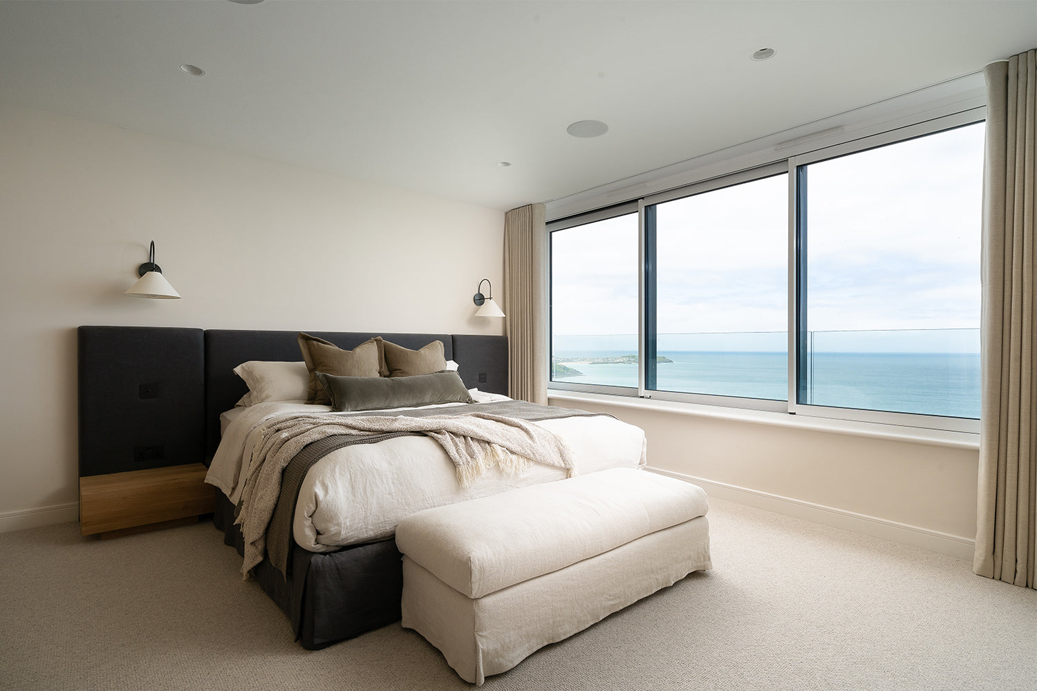 Modern coastal bedroom with panoramic ocean views, upholstered king-size bed with neutral bedding, wall-mounted sconces, soft carpet flooring, and floor-to-ceiling windows – luxury seaside interior design inspiration.