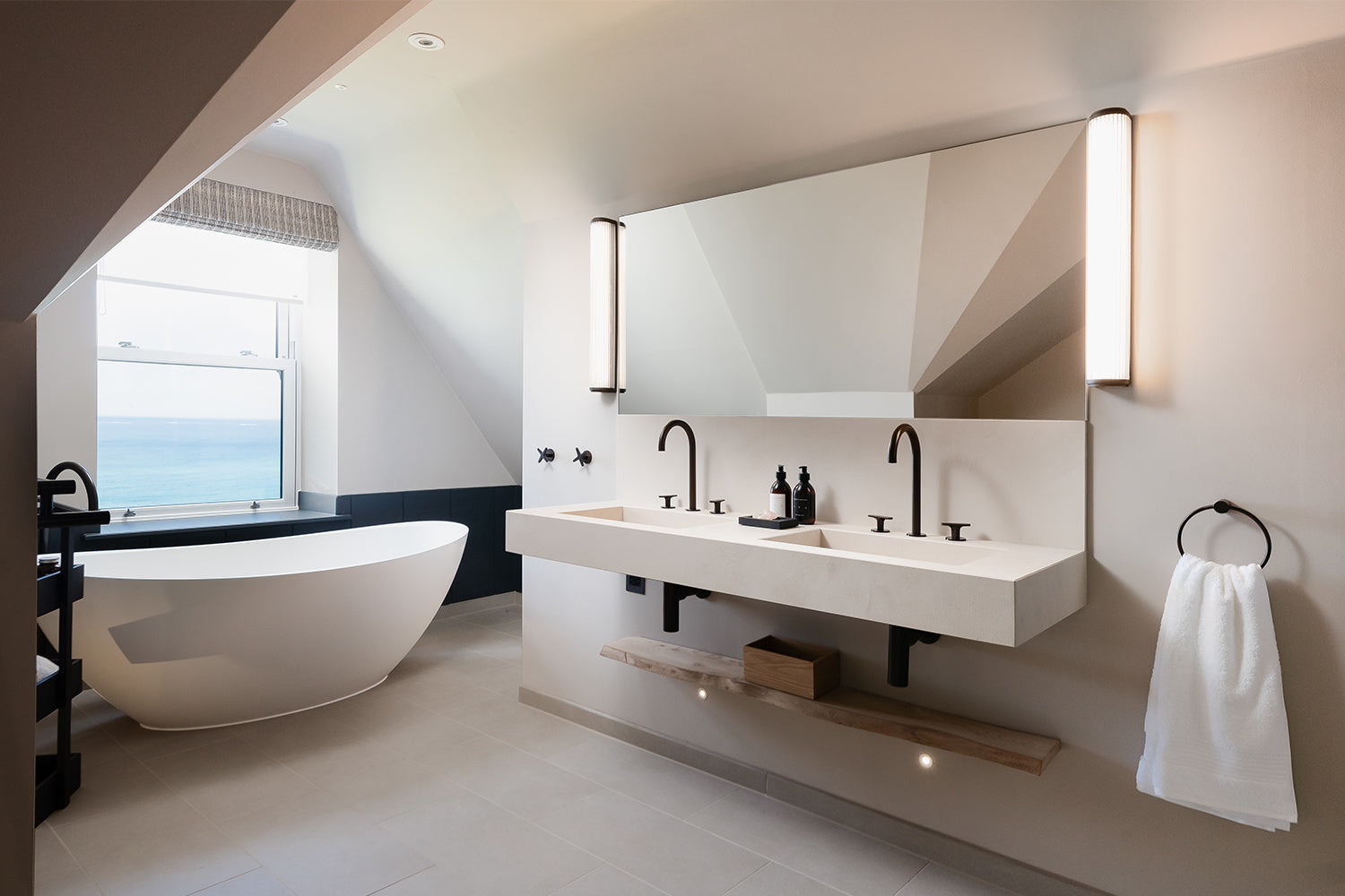 Contemporary coastal bathroom with freestanding oval bathtub, double floating vanity with matte black fixtures, minimalist wall lighting, and sea view window – luxury modern bathroom design