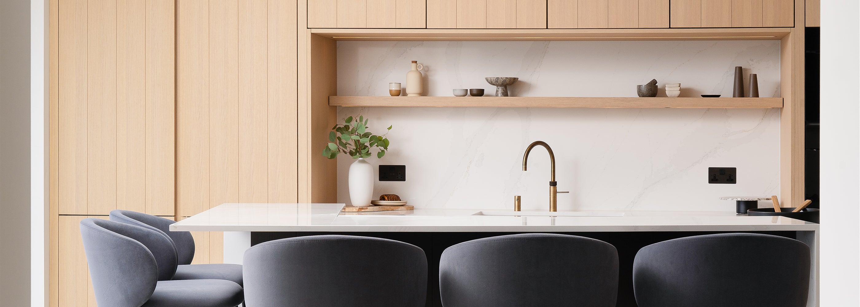 Bespoke modern kitchen with light oak cabinetry, white marble worktop, integrated open shelving, brass fixtures, and upholstered bar stools – custom kitchen design and installation by Iroka Interiors in Cornwall.
